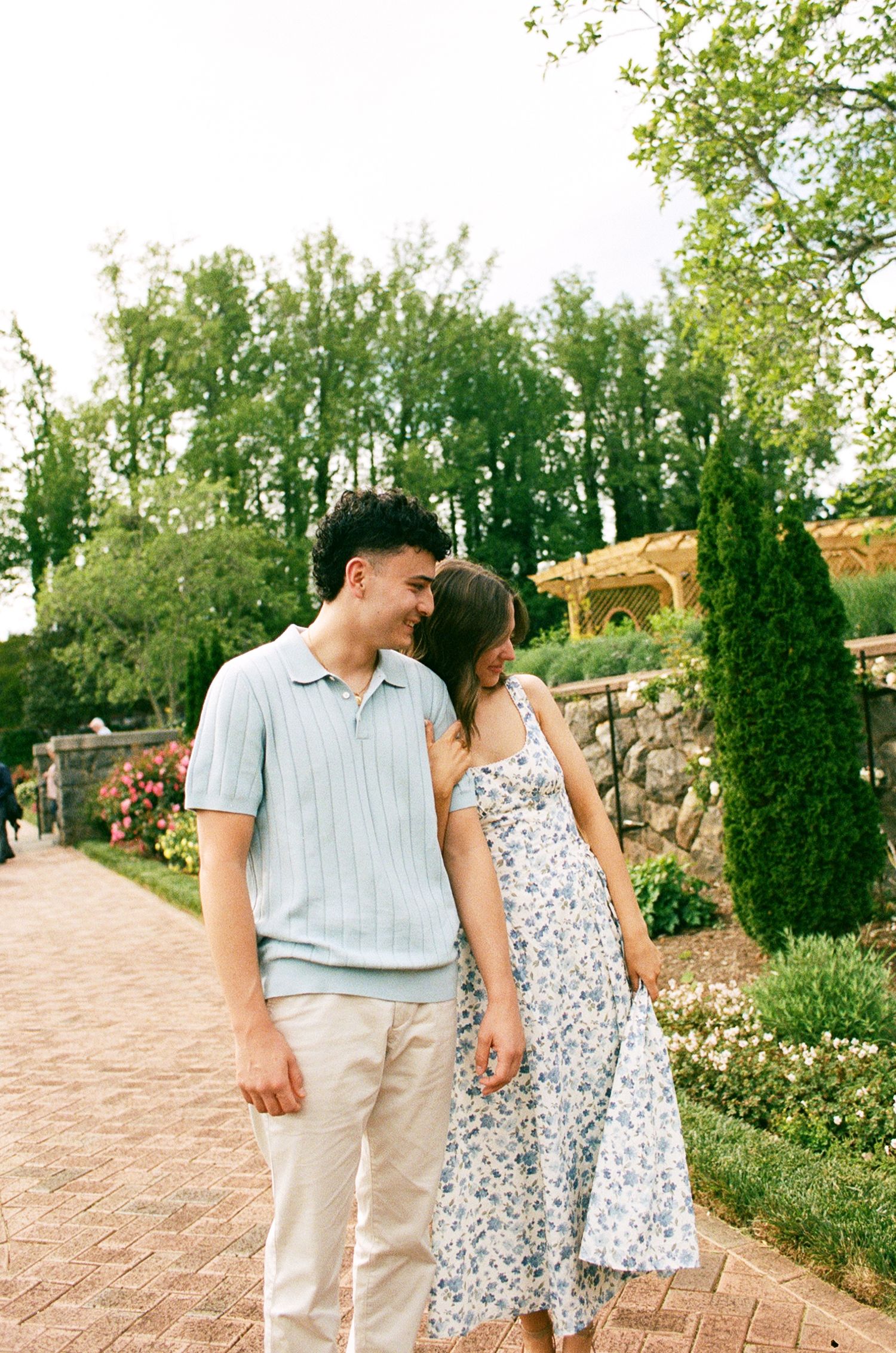 A couple walks hand-in-hand through a lush garden path, smiling at each other in casual summer attire.