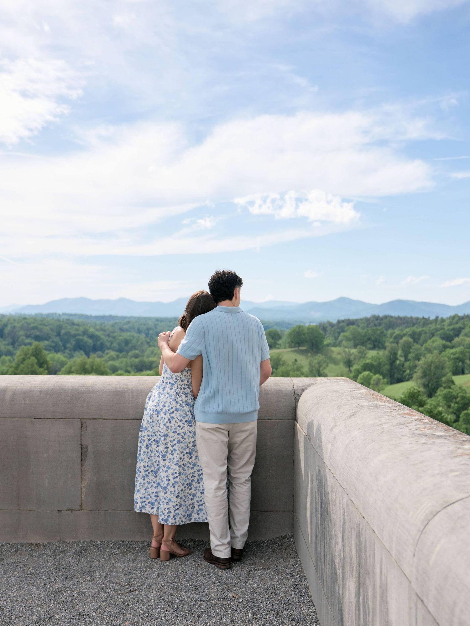 Couple embracing while overlooking scenic mountain landscape and lush green forest from a concrete terrace on a sunny day.