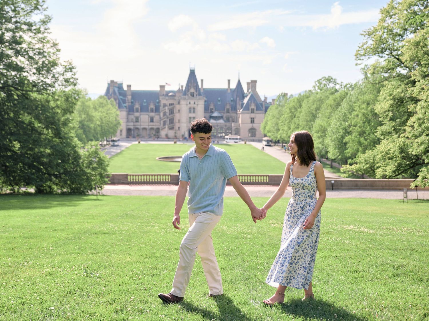 Couple holding hands on green lawn with Biltmore Estate in background.