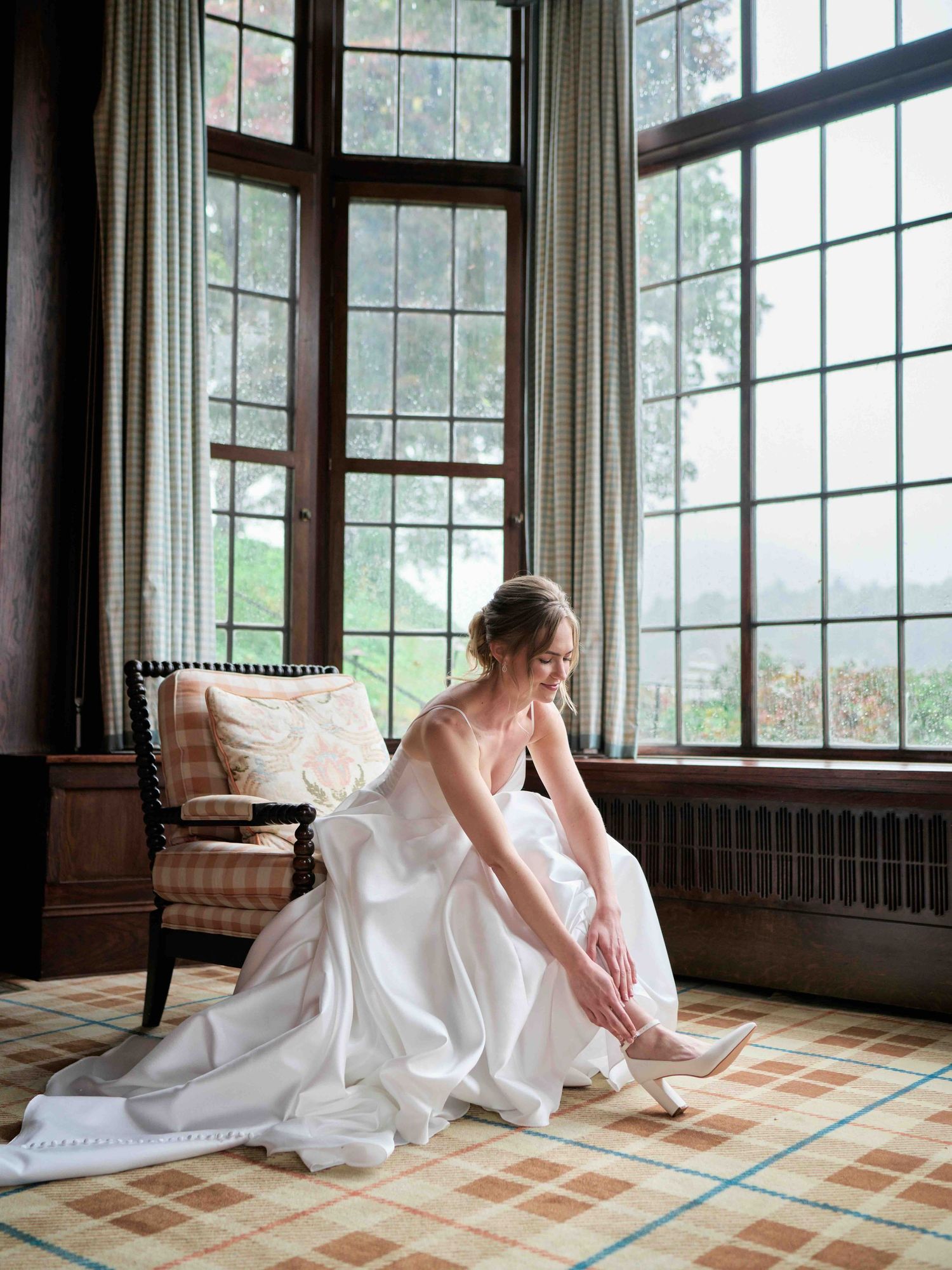 Bride in white gown adjusting her heel by large windows in an elegant room with plaid carpet.