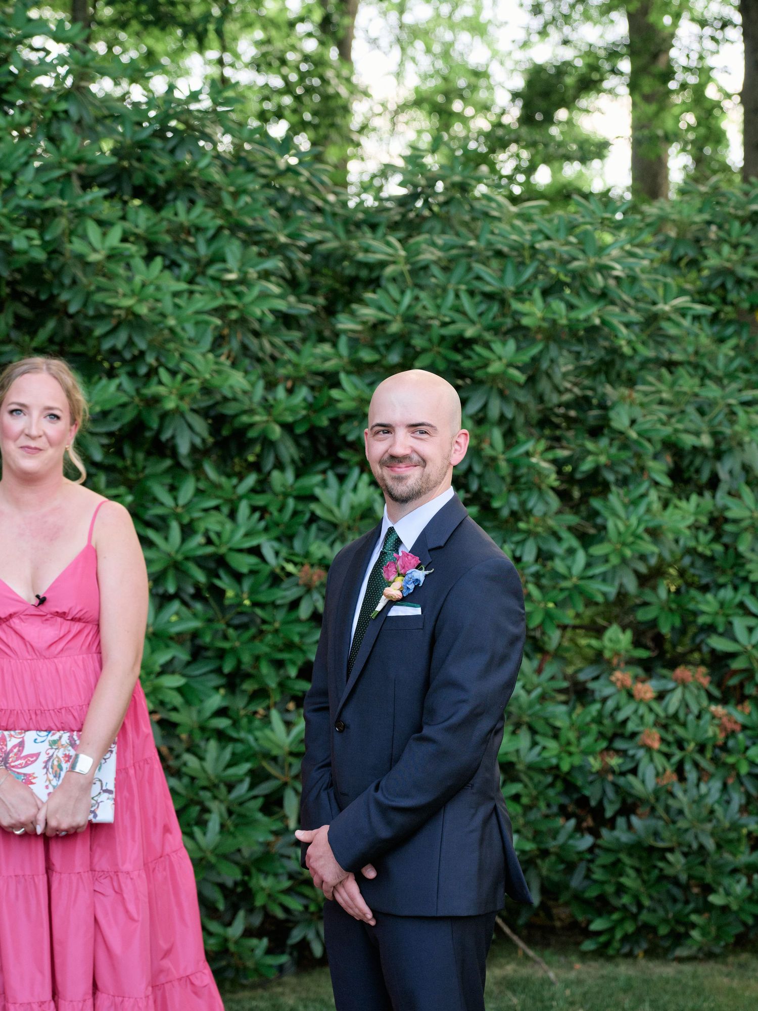Formal event attendees pose against green hedge backdrop at outdoor venue.