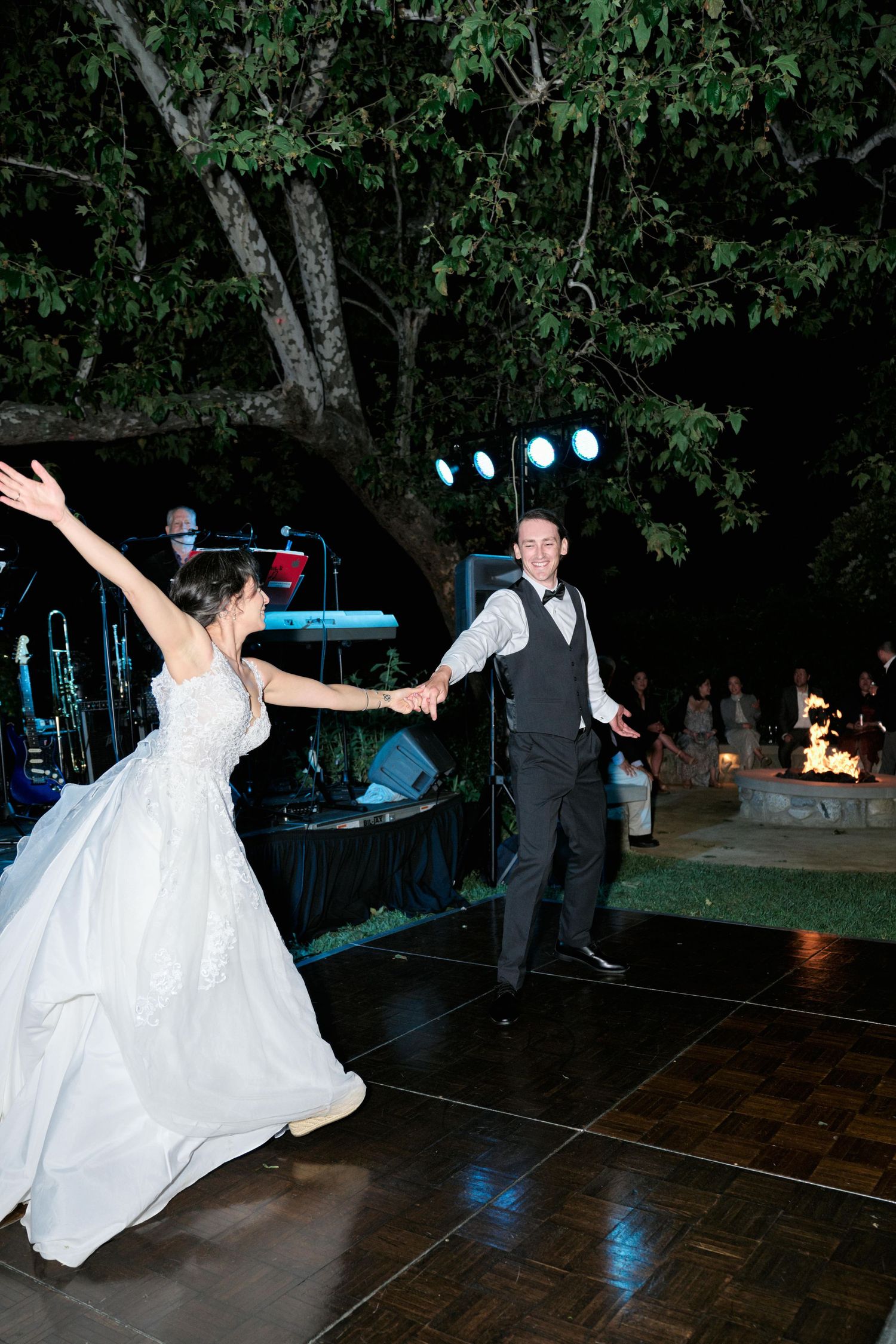 A newlywed couple dances energetically on an outdoor dance floor under tree branches and stage lights.