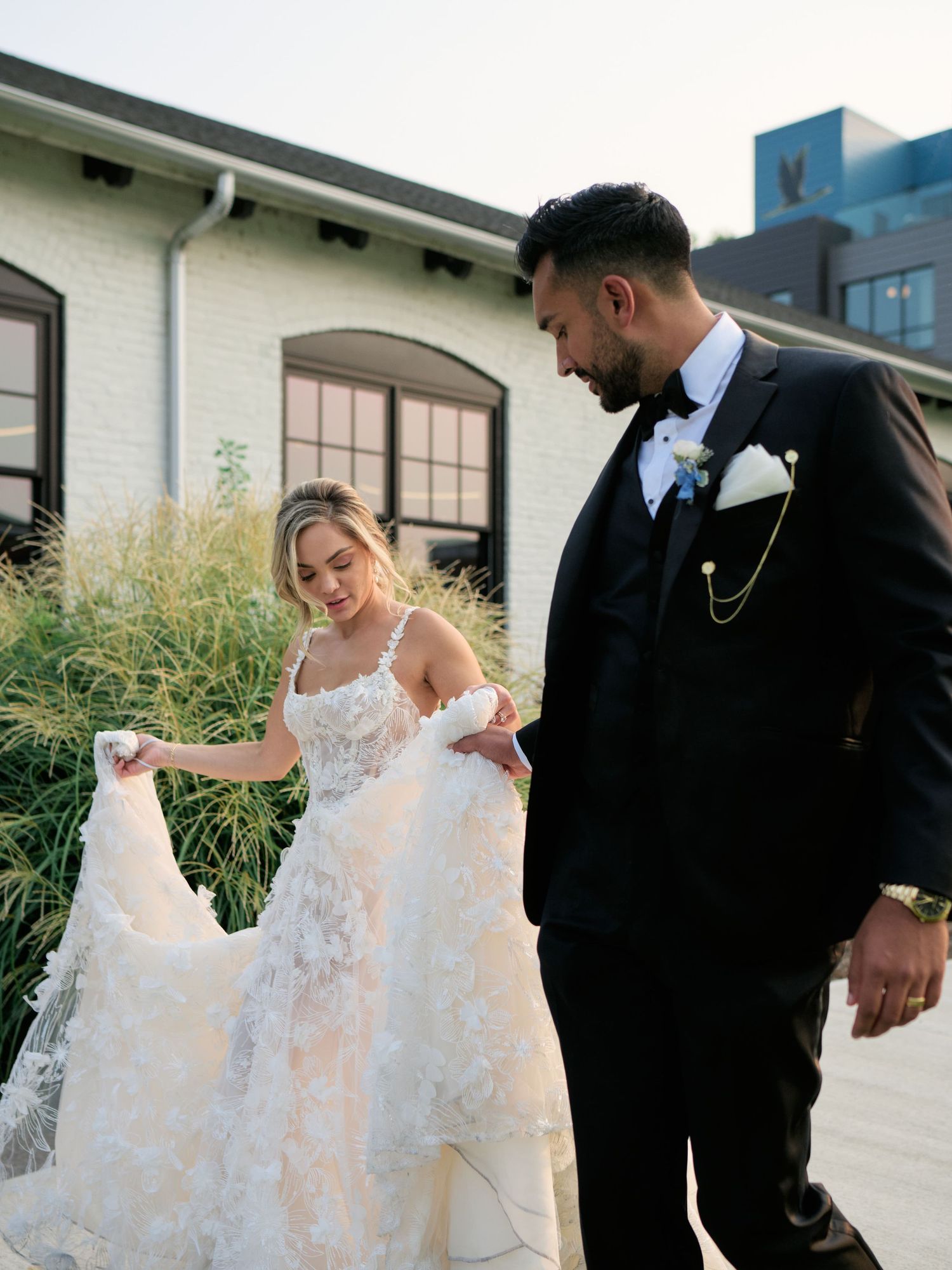 A couple in wedding attire outdoors, with the bride holding up her lace dress train while wearing a white gown.