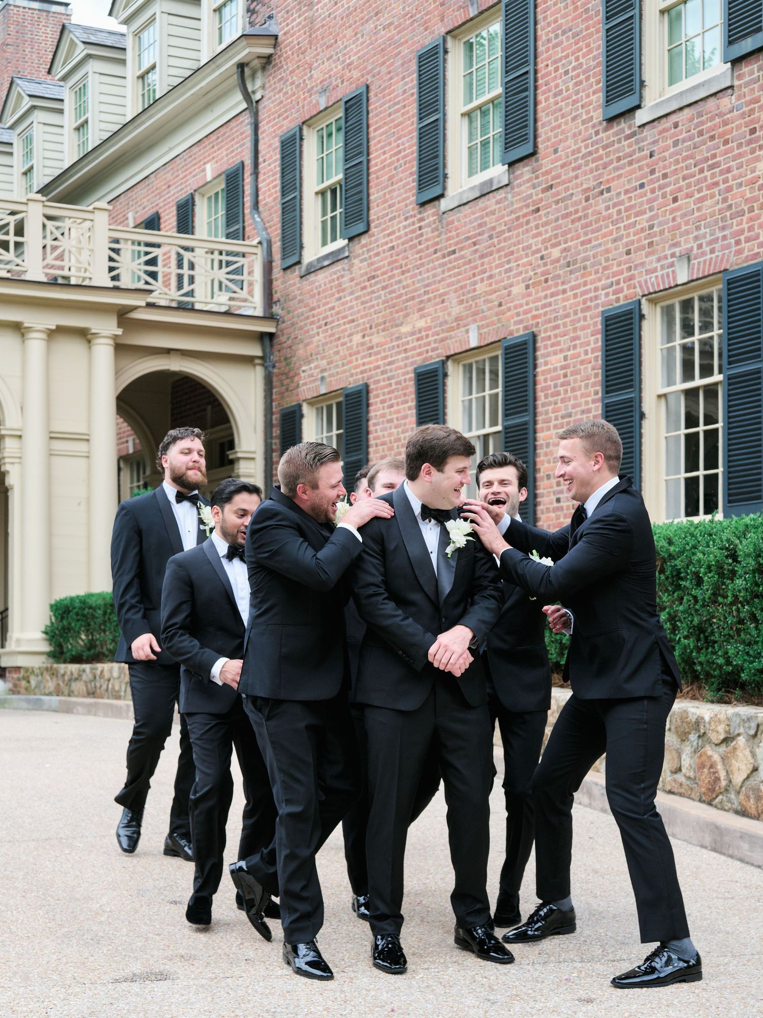 Groomsmen in black tuxedos share candid laughing moment outside historic brick building.