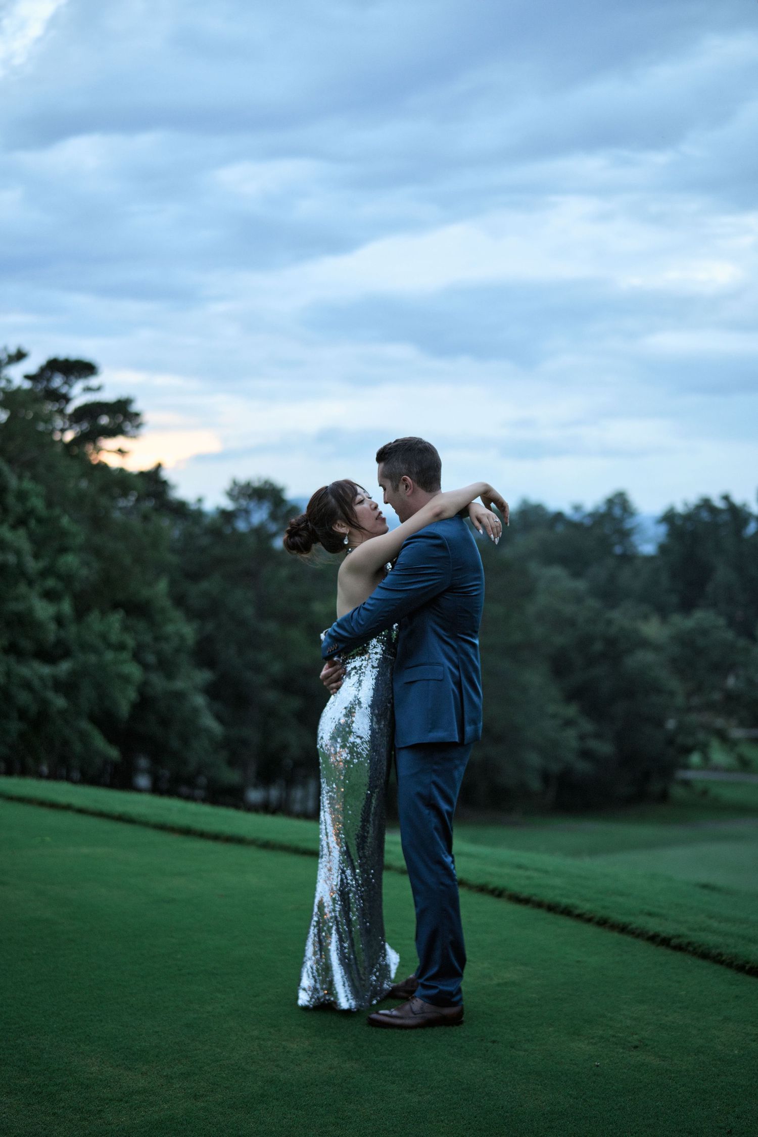 A romantic wedding couple embraces on a scenic golf course at dusk with lush greenery in the background.