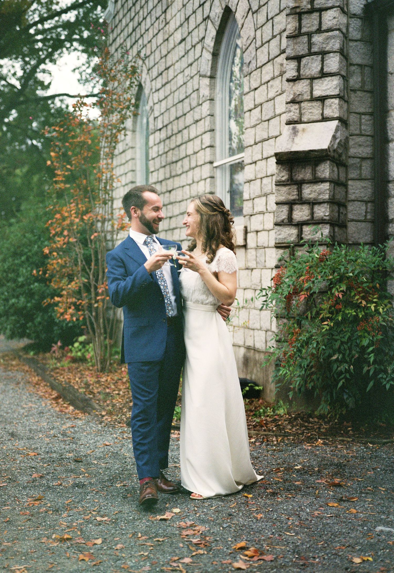 A couple shares a toast outside a historic stone church building surrounded by autumn foliage.
