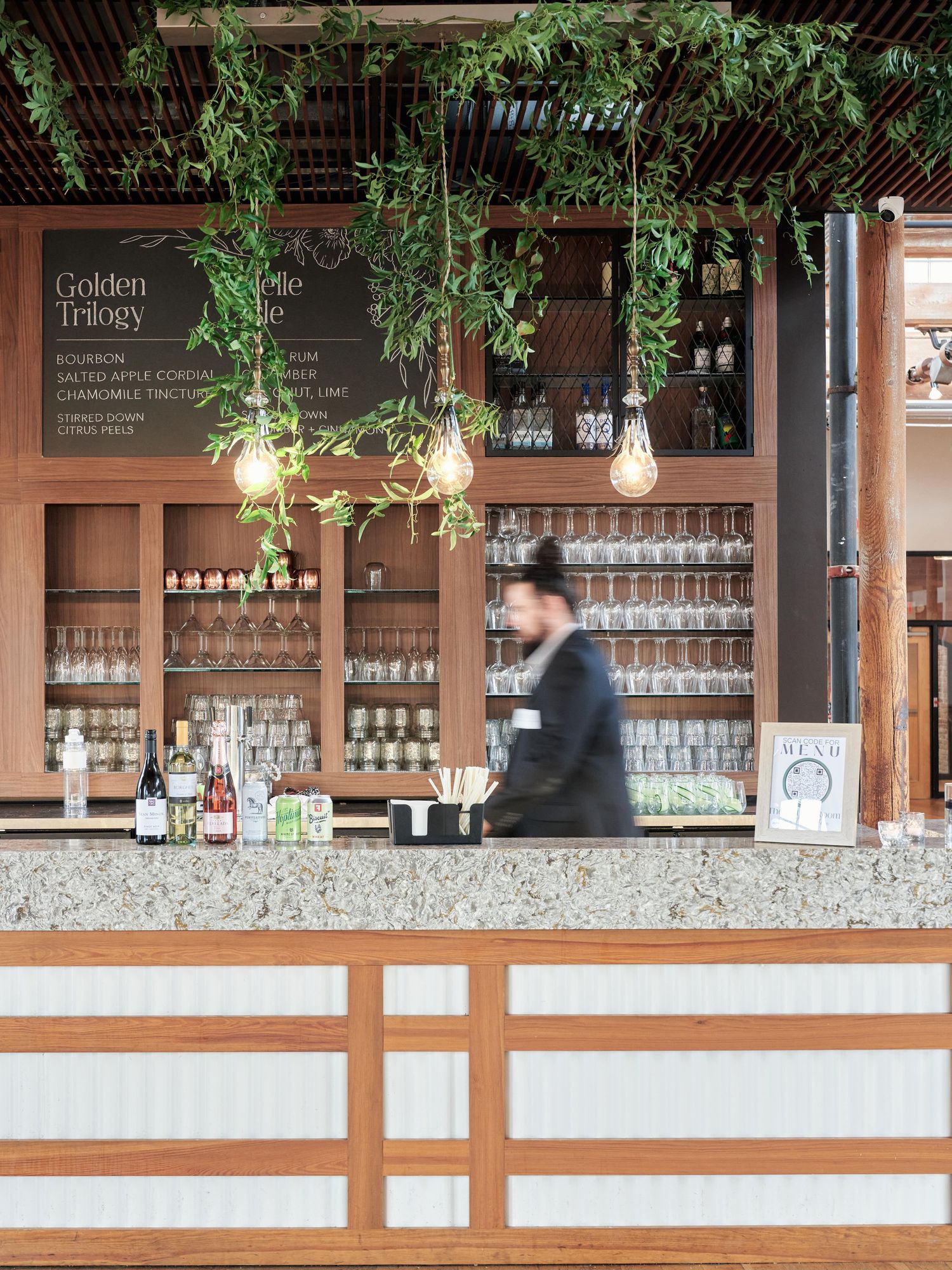 Rustic bar with hanging lights and greenery, featuring wooden shelving and blurred bartender.
