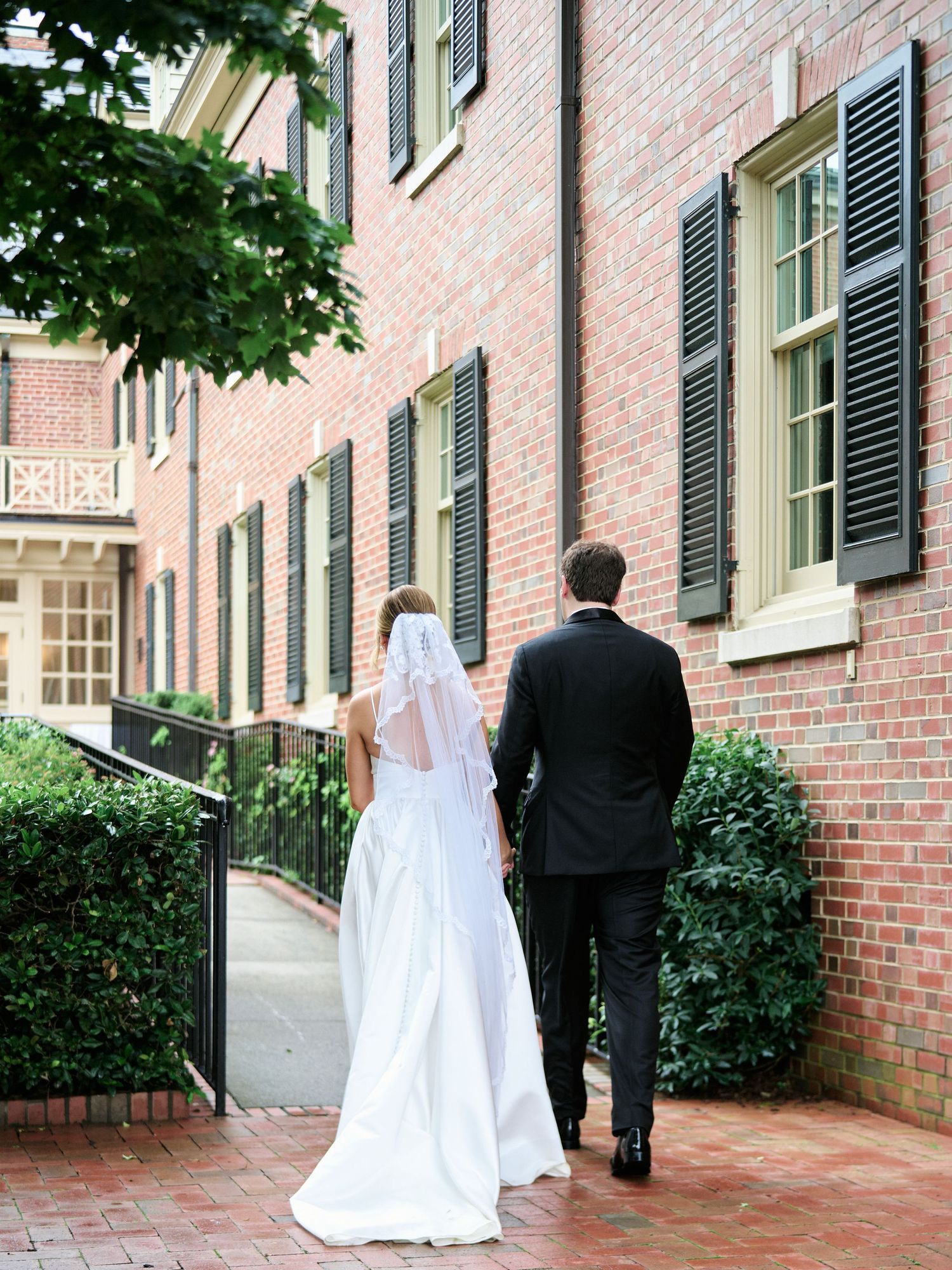 A bride and groom walk along a brick path beside a historic building exterior.