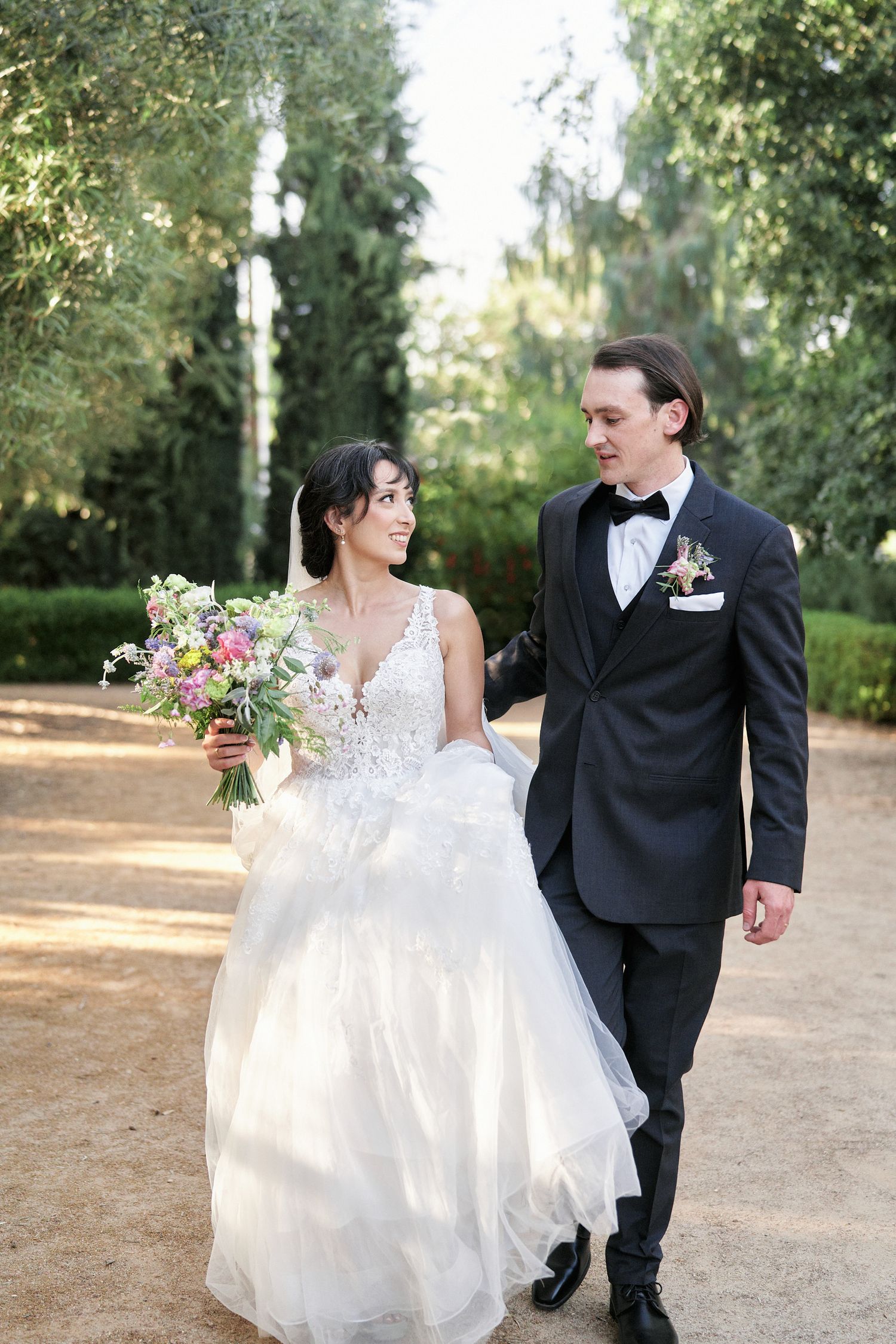A couple in formal wedding attire walks together on a garden path while sharing a joyful moment.