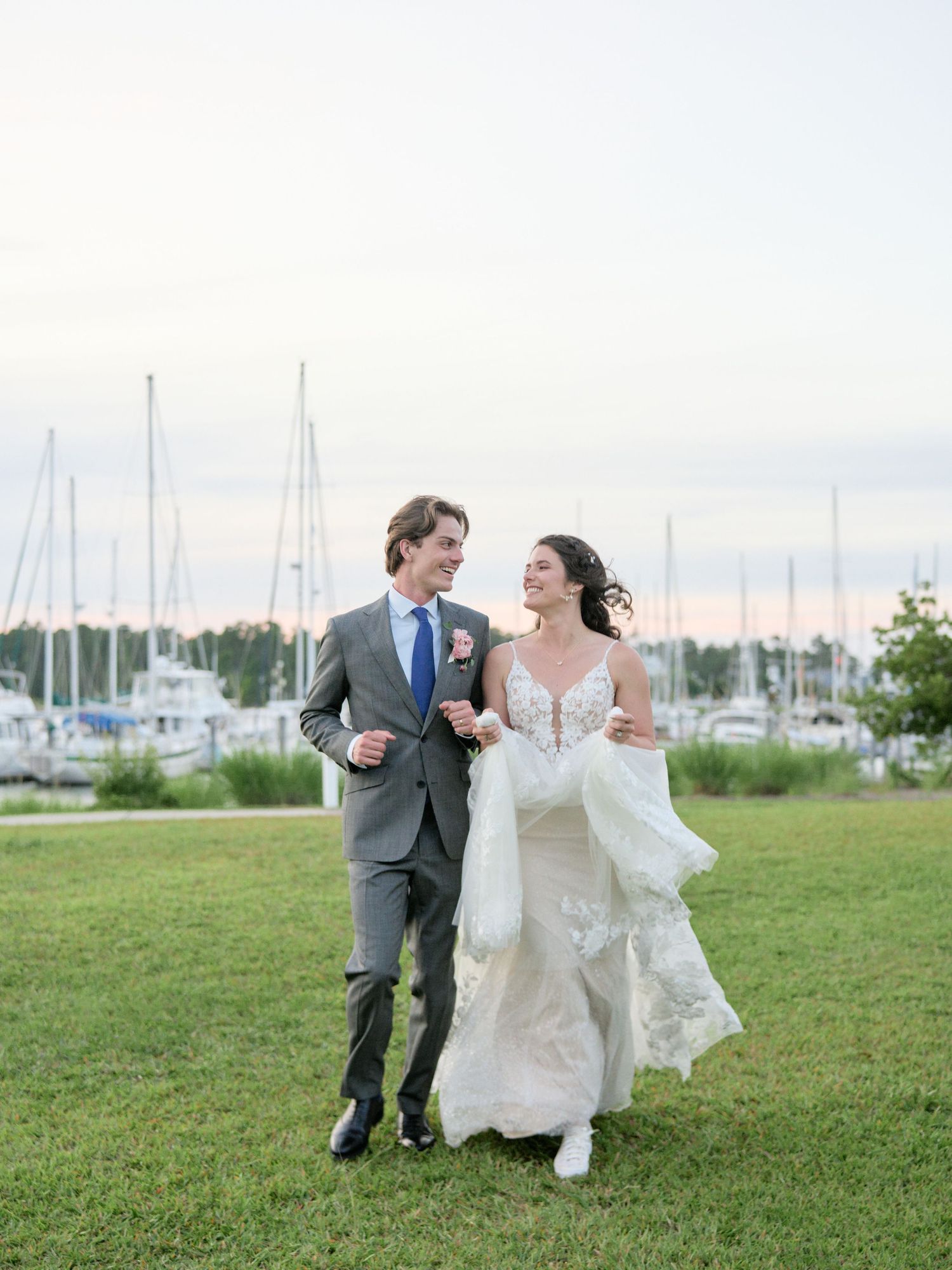 A newlywed couple walking together on grass with sailboats docked at a marina in the background.