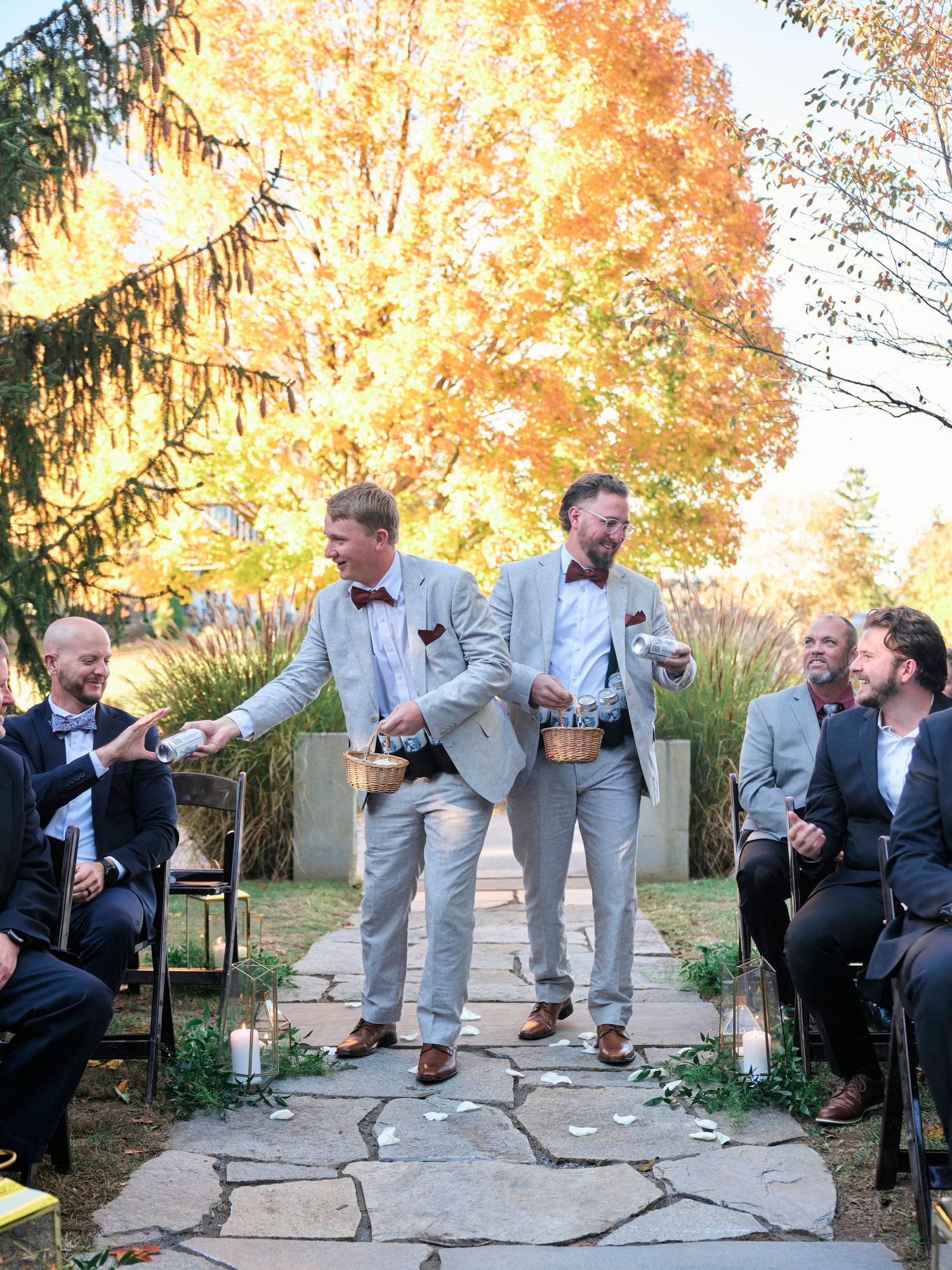 Wedding party members share a lighthearted moment on a stone path with vibrant autumn trees in the background.