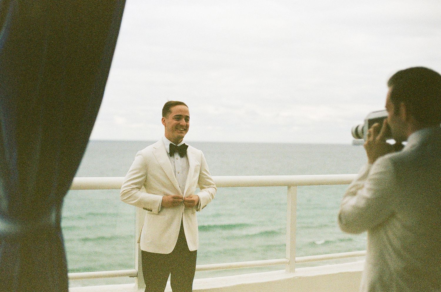 A person in a cream jacket poses while being photographed on a balcony overlooking the ocean.