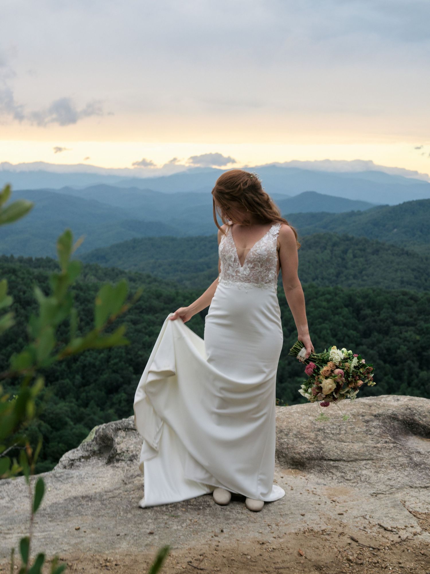 A bride in a fitted white dress stands on a mountain cliff edge holding a bouquet at sunset.