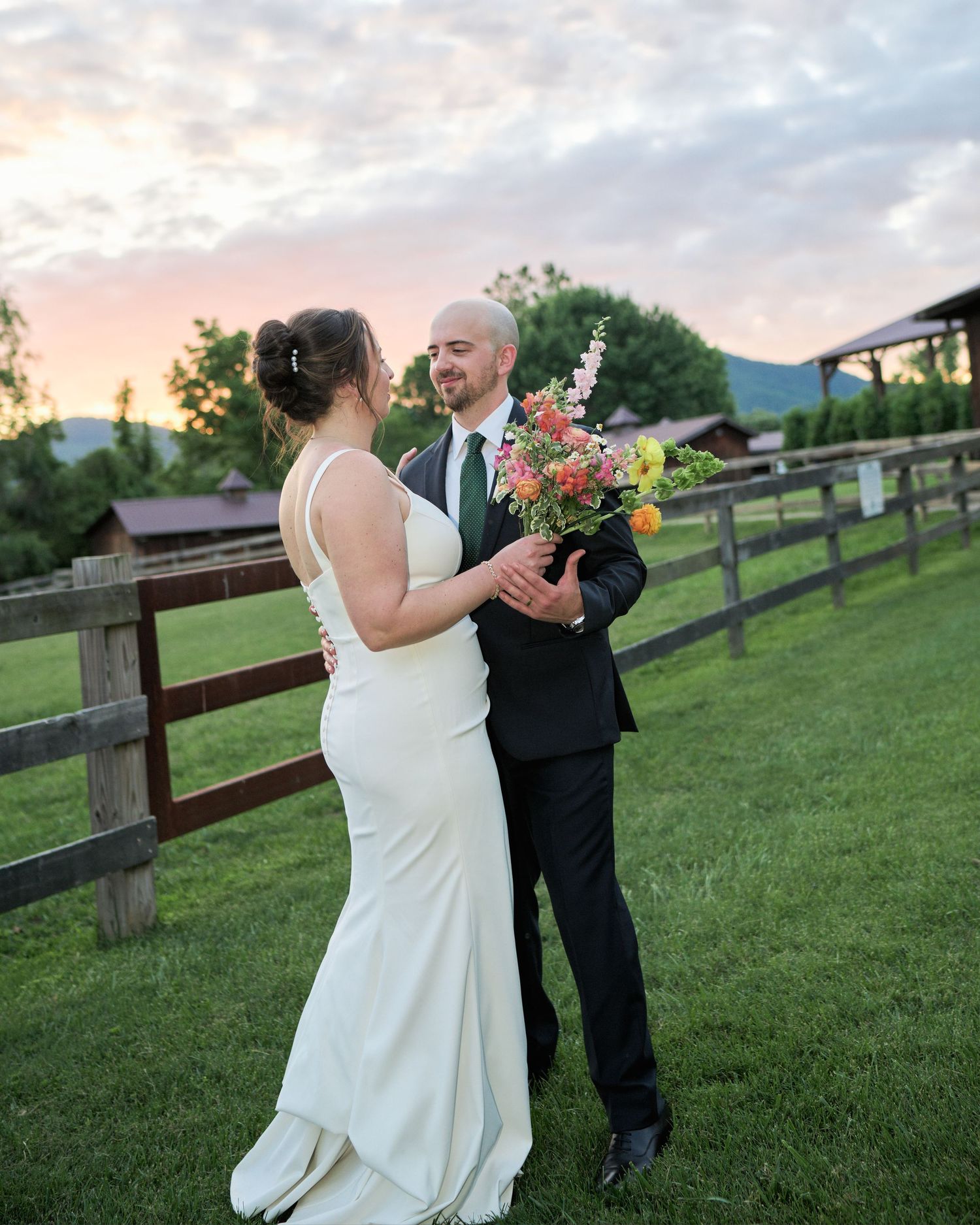 A couple in wedding attire embrace at sunset on a rustic farm property with wooden fences and pink flowers.