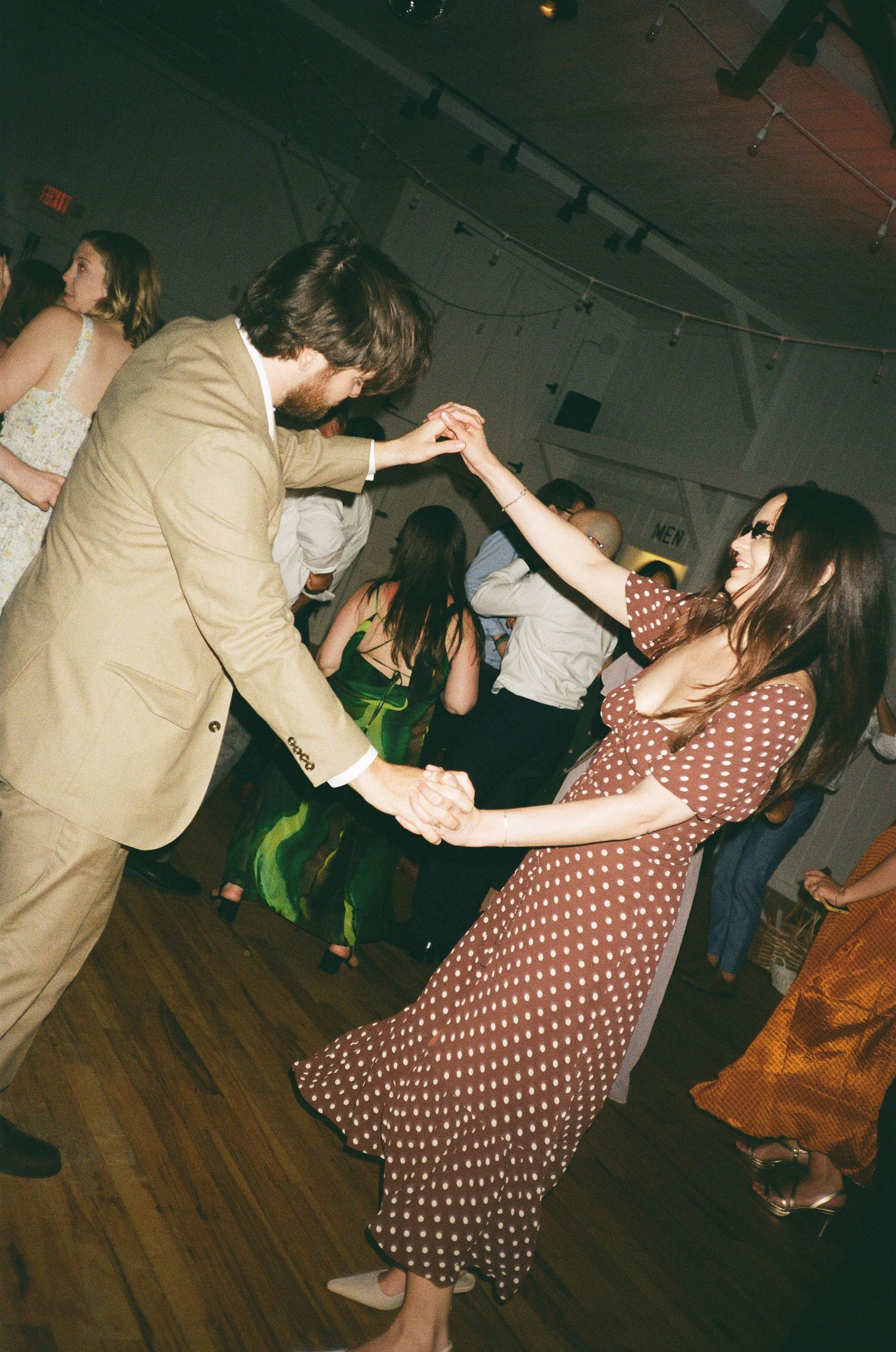 Couple dancing together at a wedding reception, man in tan suit, woman in brown polka dot dress, on a wooden dance floor.