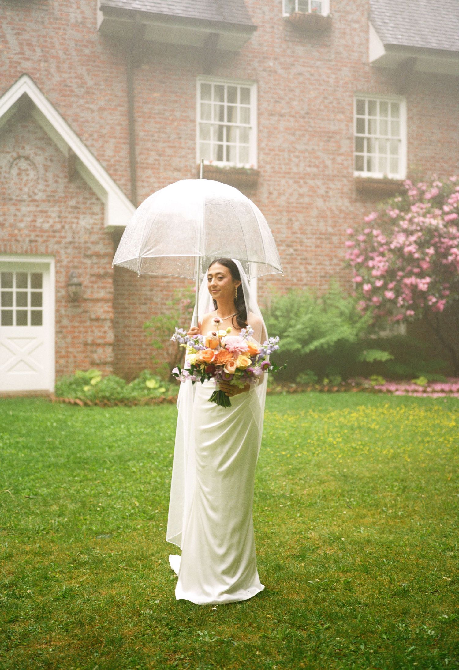 Bride in white gown holding clear umbrella and colorful bouquet standing on green lawn before brick mansion.