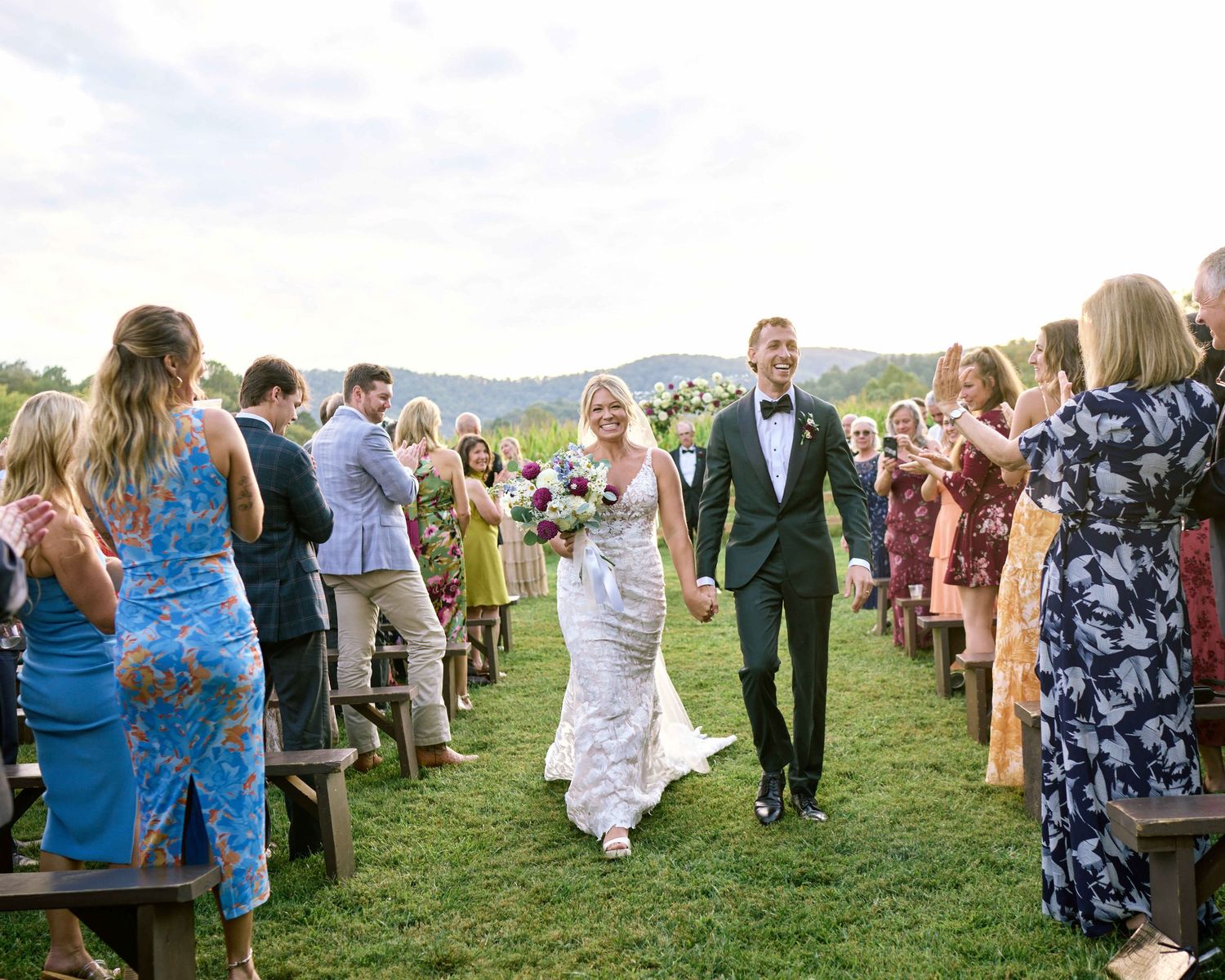 Wedding guests mingle and socialize on a grassy hillside during an outdoor reception celebration.