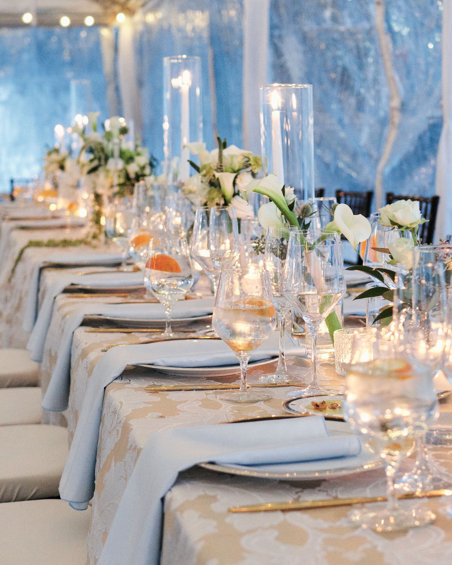 Elegant wedding reception table with white floral centerpieces, crystal glassware, and gold accents under a blue-lit tent.