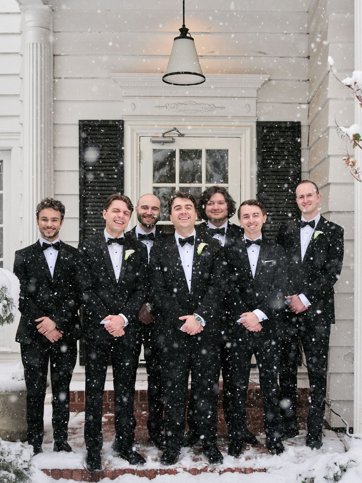 Seven groomsmen in black tuxedos pose on a snowy porch during a winter wedding celebration.