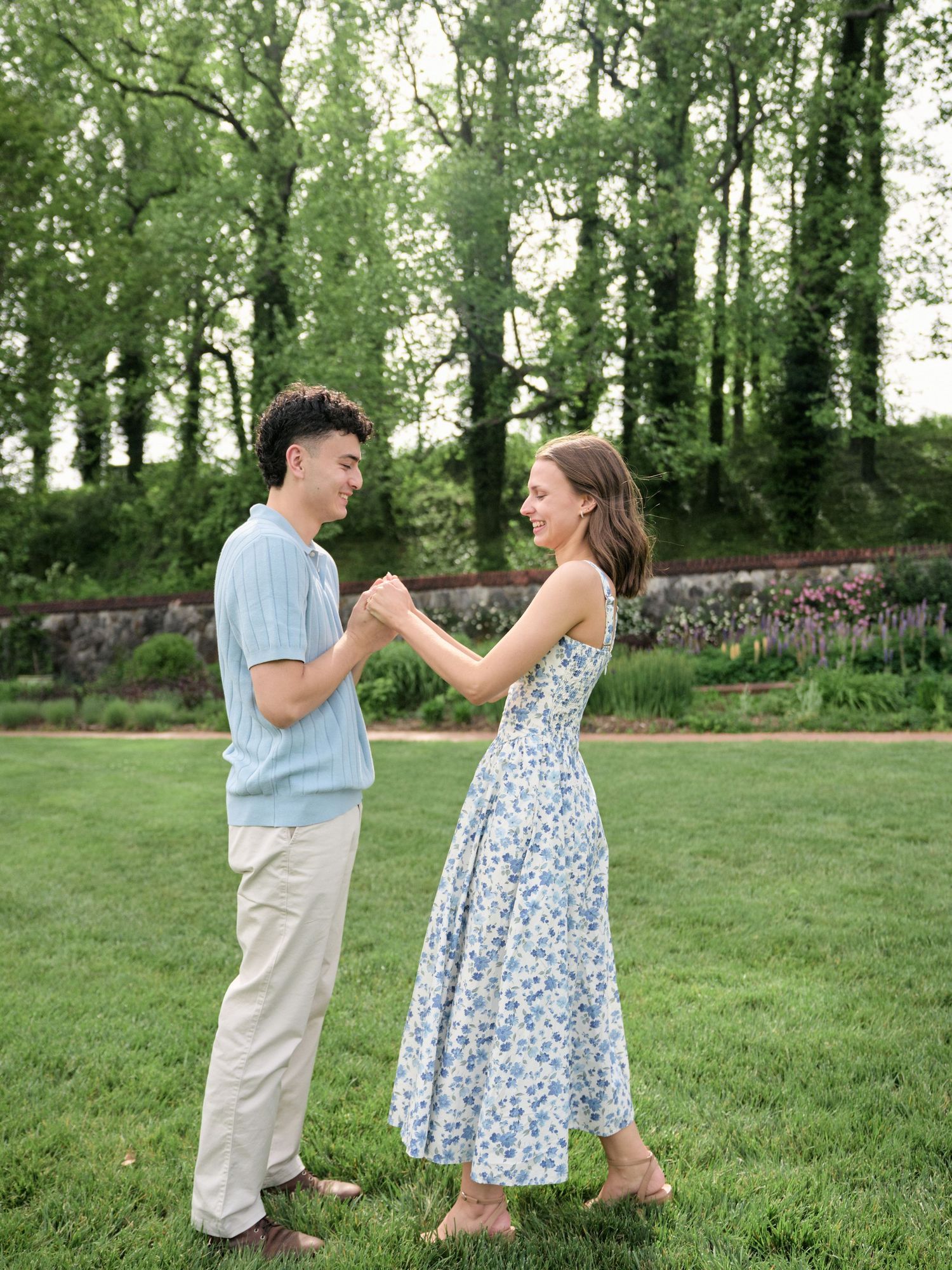 A couple dances barefoot on a green lawn, the woman in a floral dress and the man in a blue shirt, surrounded by lush trees.