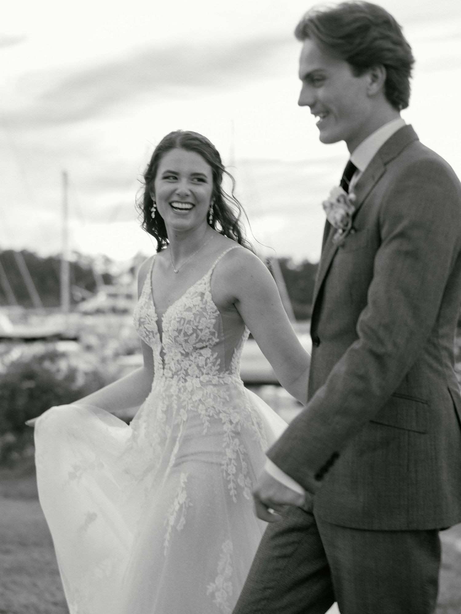 Black and white photo of a smiling bride in lace gown holding hands with groom in suit at waterfront wedding.