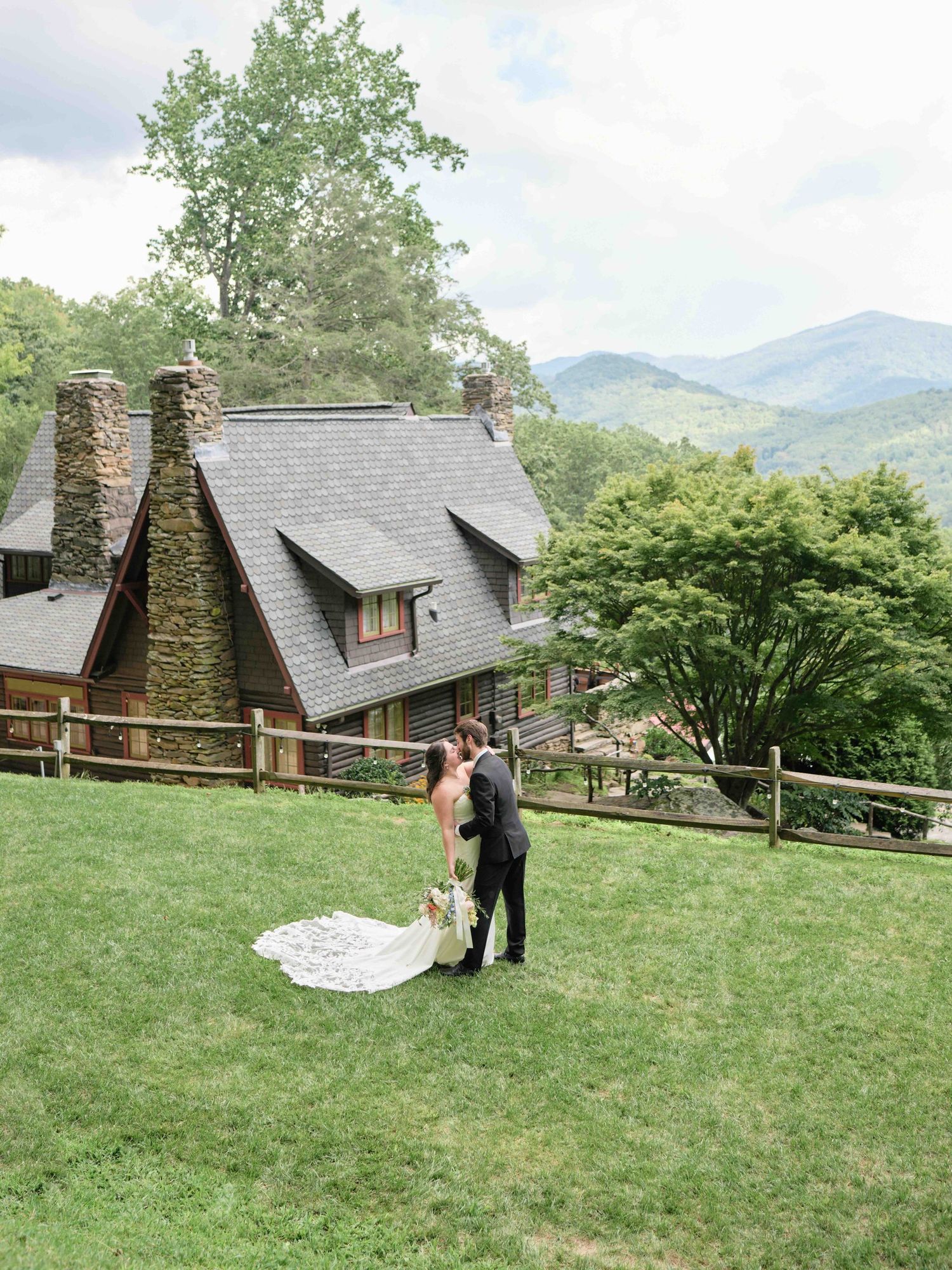 Bride and groom share a romantic kiss on a green hillside with a rustic stone cabin and misty mountain backdrop.