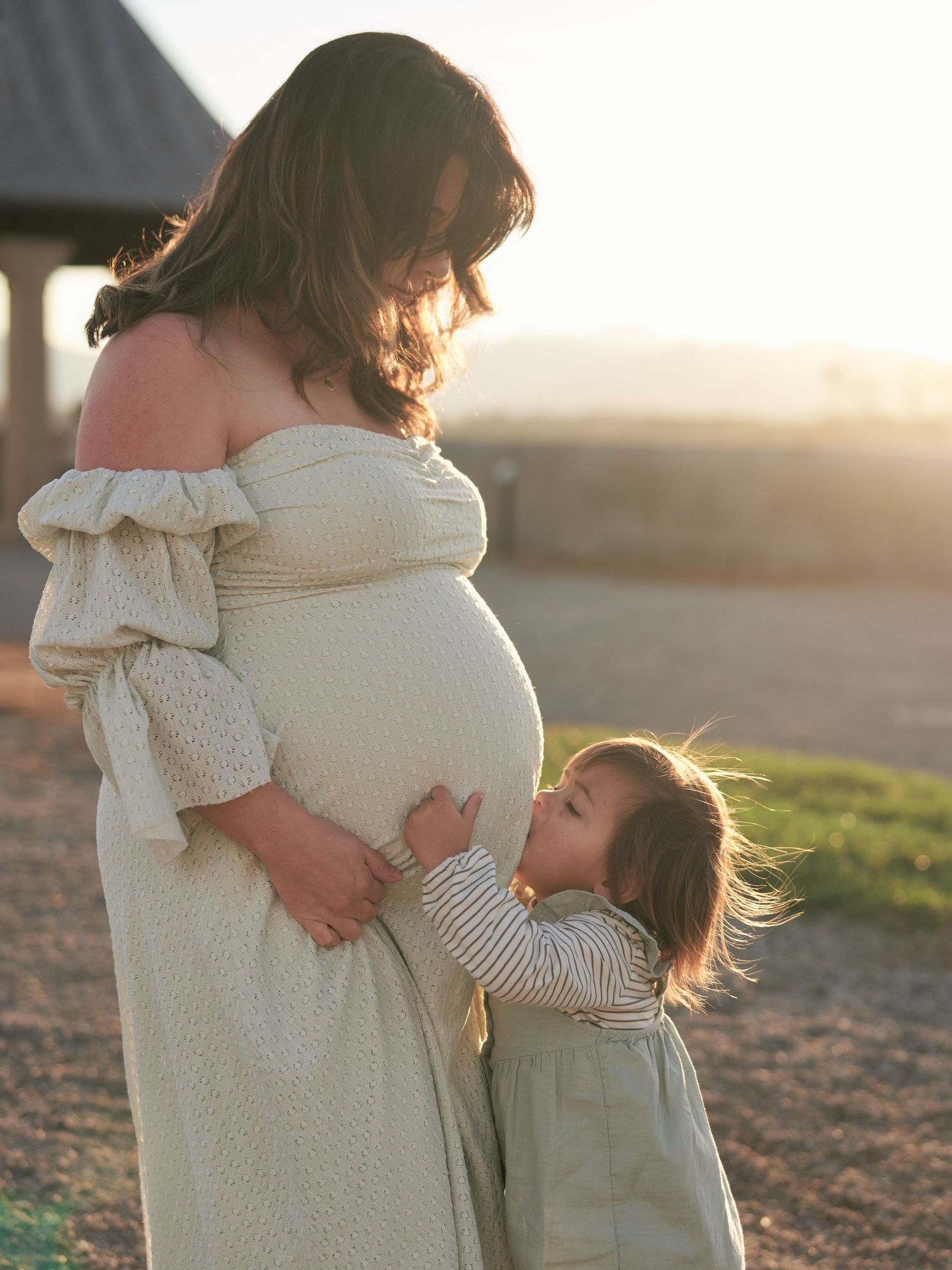 Pregnant woman in off-shoulder dress with toddler hugging her baby bump outdoors at golden hour.