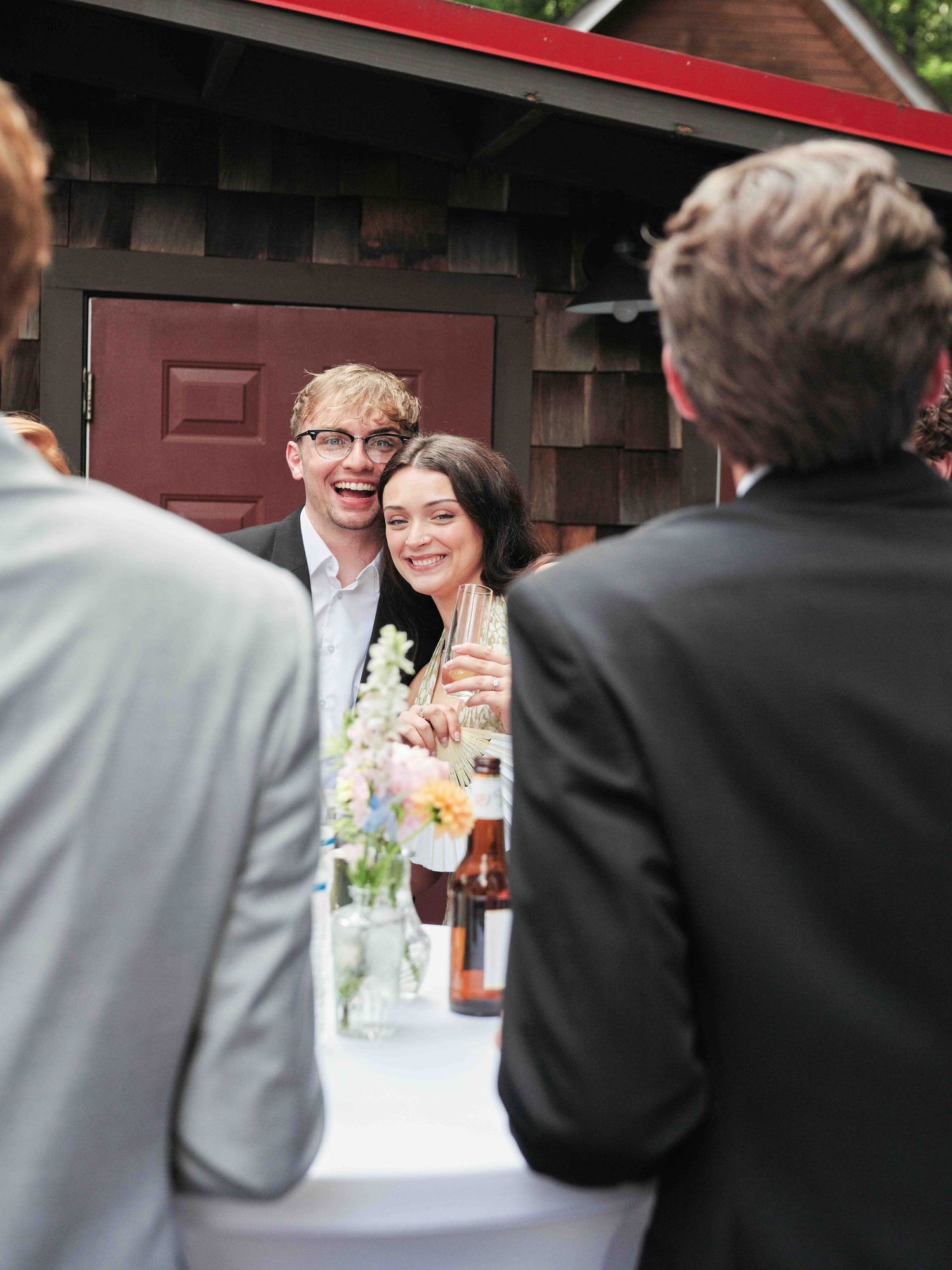 Smiling couple at an outdoor wedding reception standing near a flower-decorated table by a rustic wooden building.