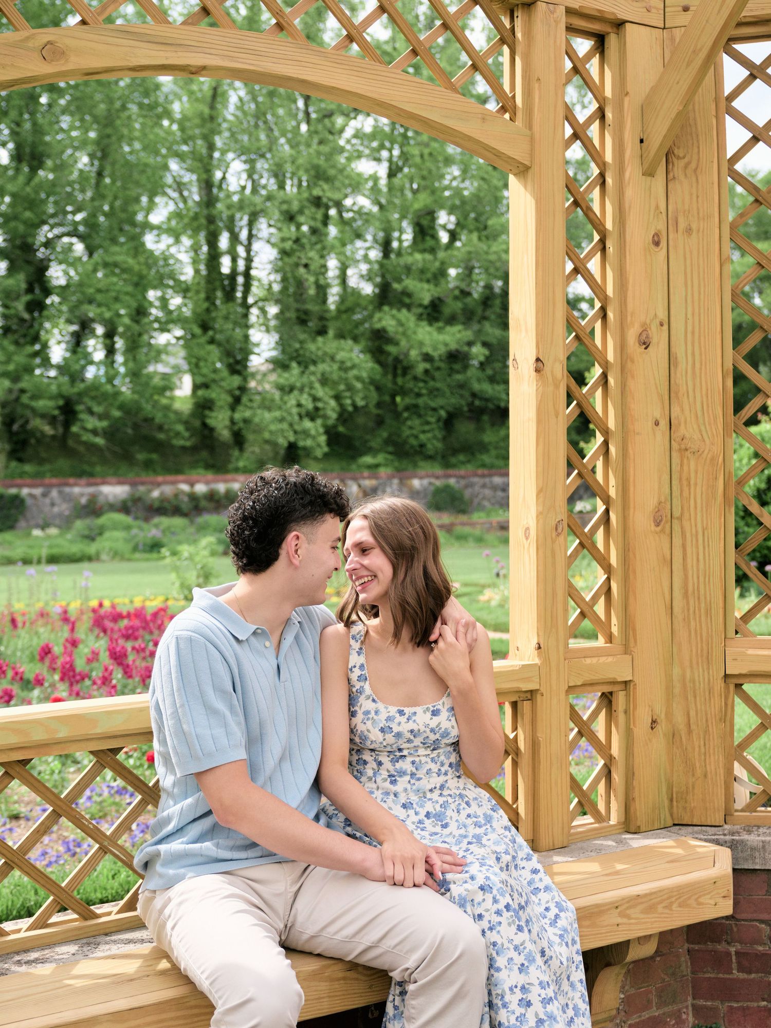A couple sits laughing together under a wooden garden arbor surrounded by lush greenery and colorful flowers.