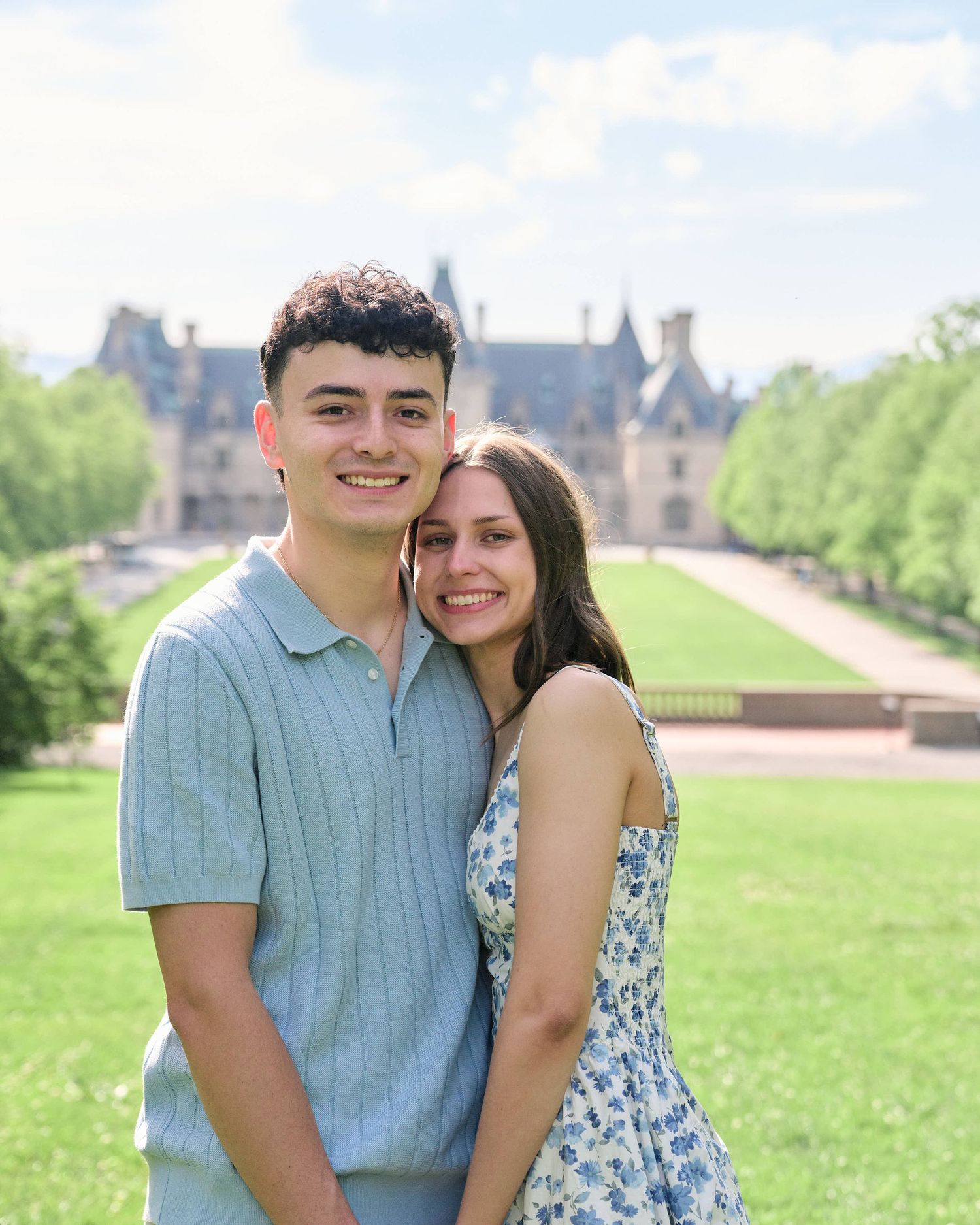 A young couple smiles together on a green lawn with a grand château in the background on a sunny day.