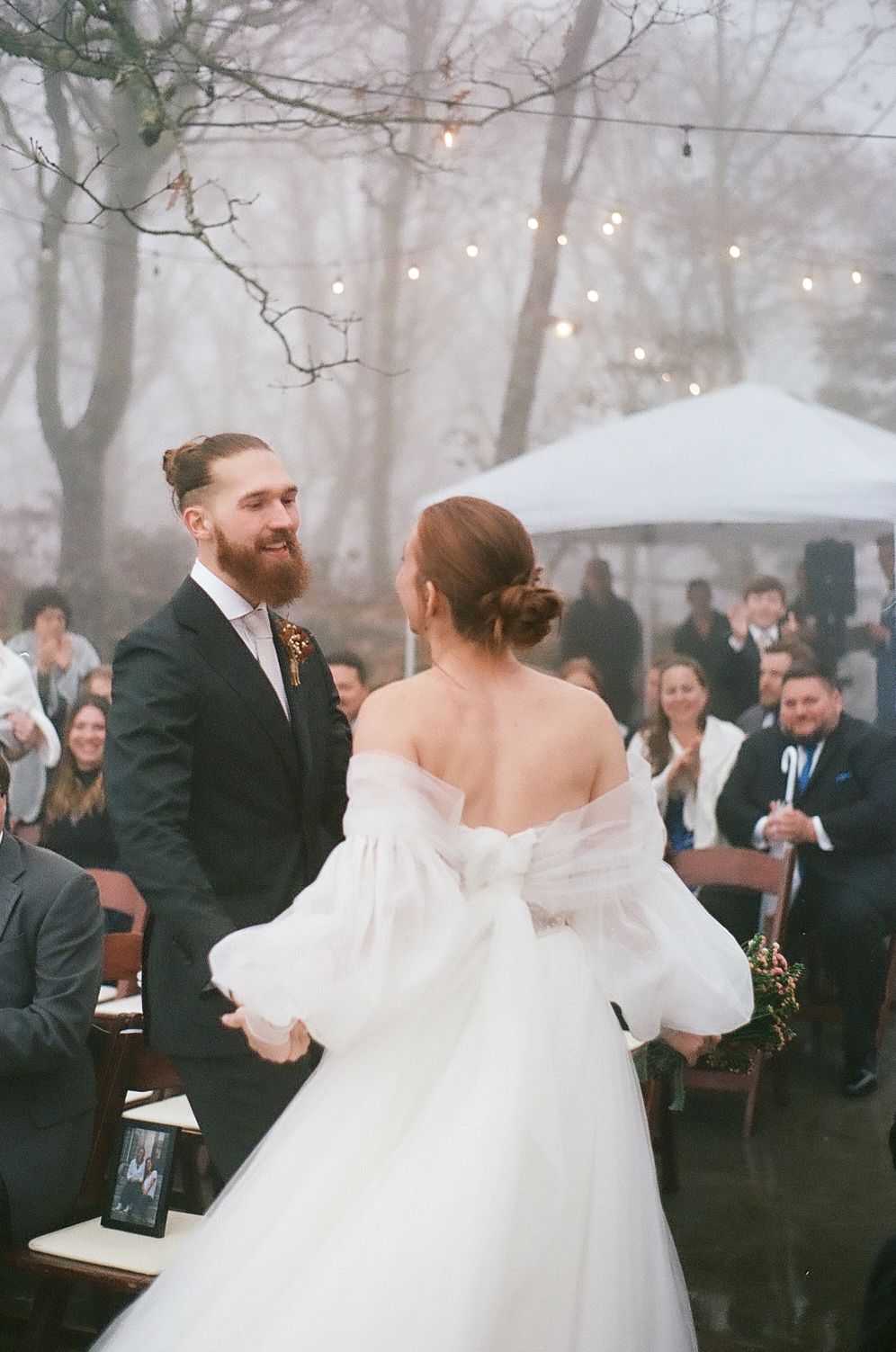 An outdoor wedding ceremony in a foggy forest with string lights and white tent.