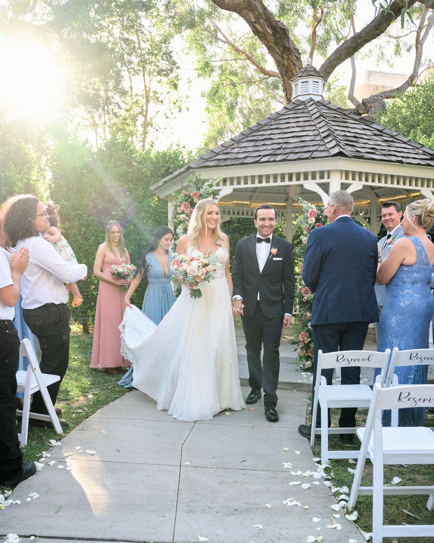 Newlyweds exit their garden ceremony while guests cheer under golden sunlight.