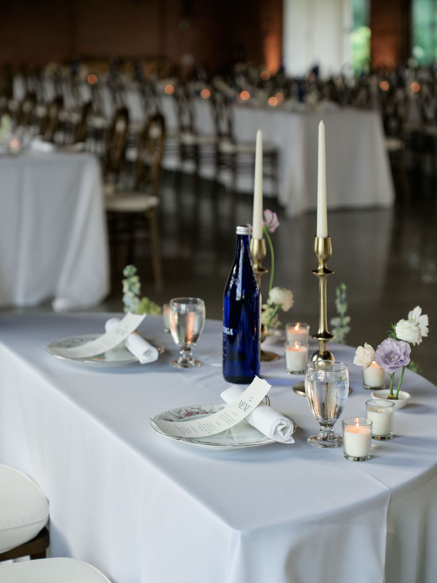 Elegant wedding reception table setting with white linens, blue glass bottles, tall candles and delicate floral arrangements.