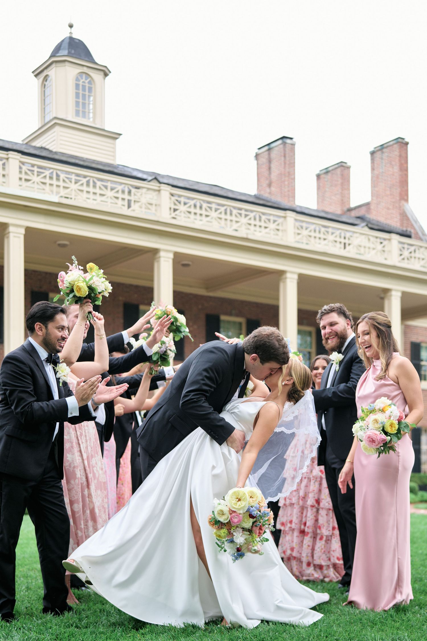 Wedding party celebrates with fun poses under columned portico of historic mansion.