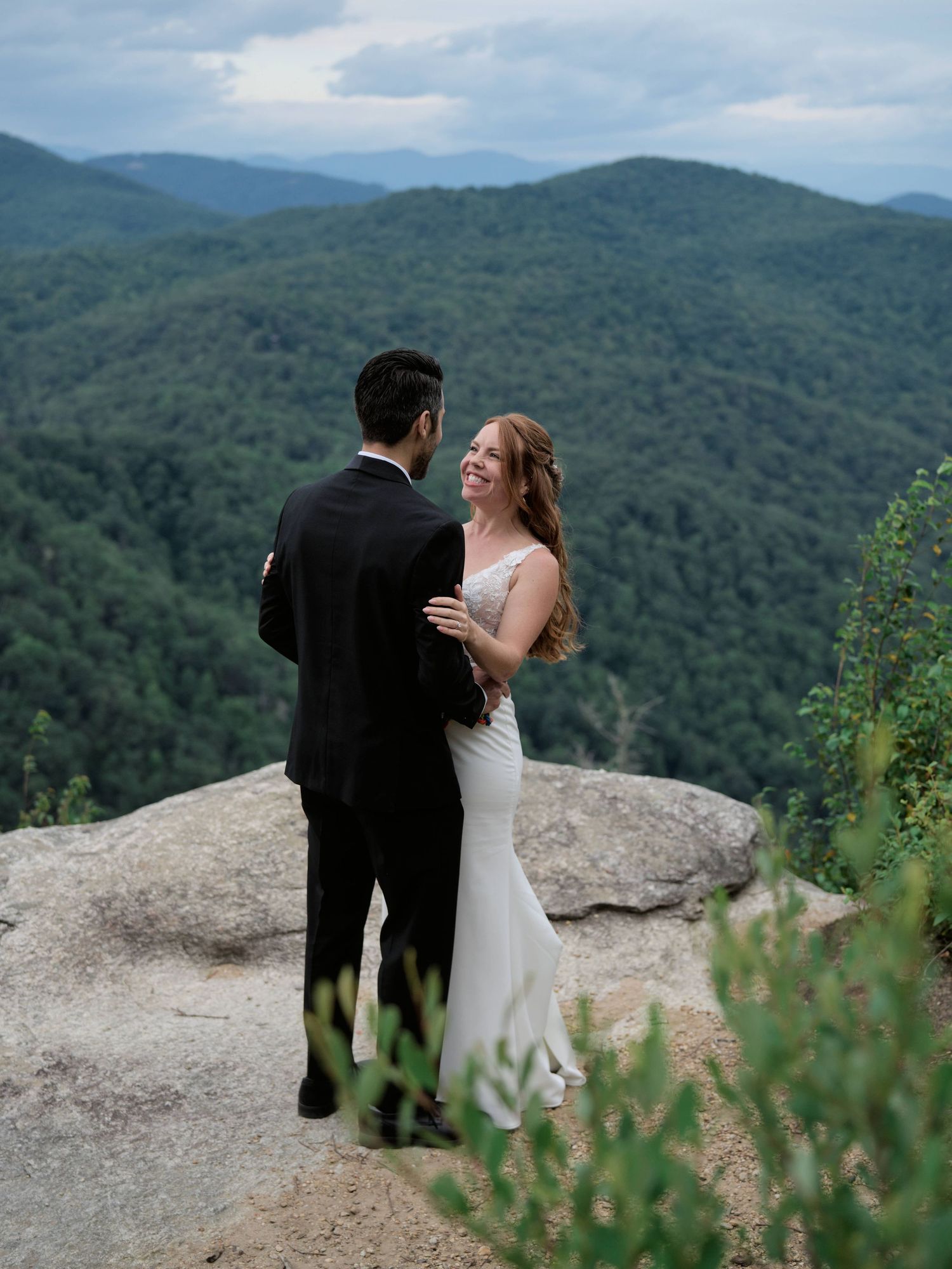 A couple shares a romantic moment on a mountain overlook with rolling green hills stretching into the misty distance.