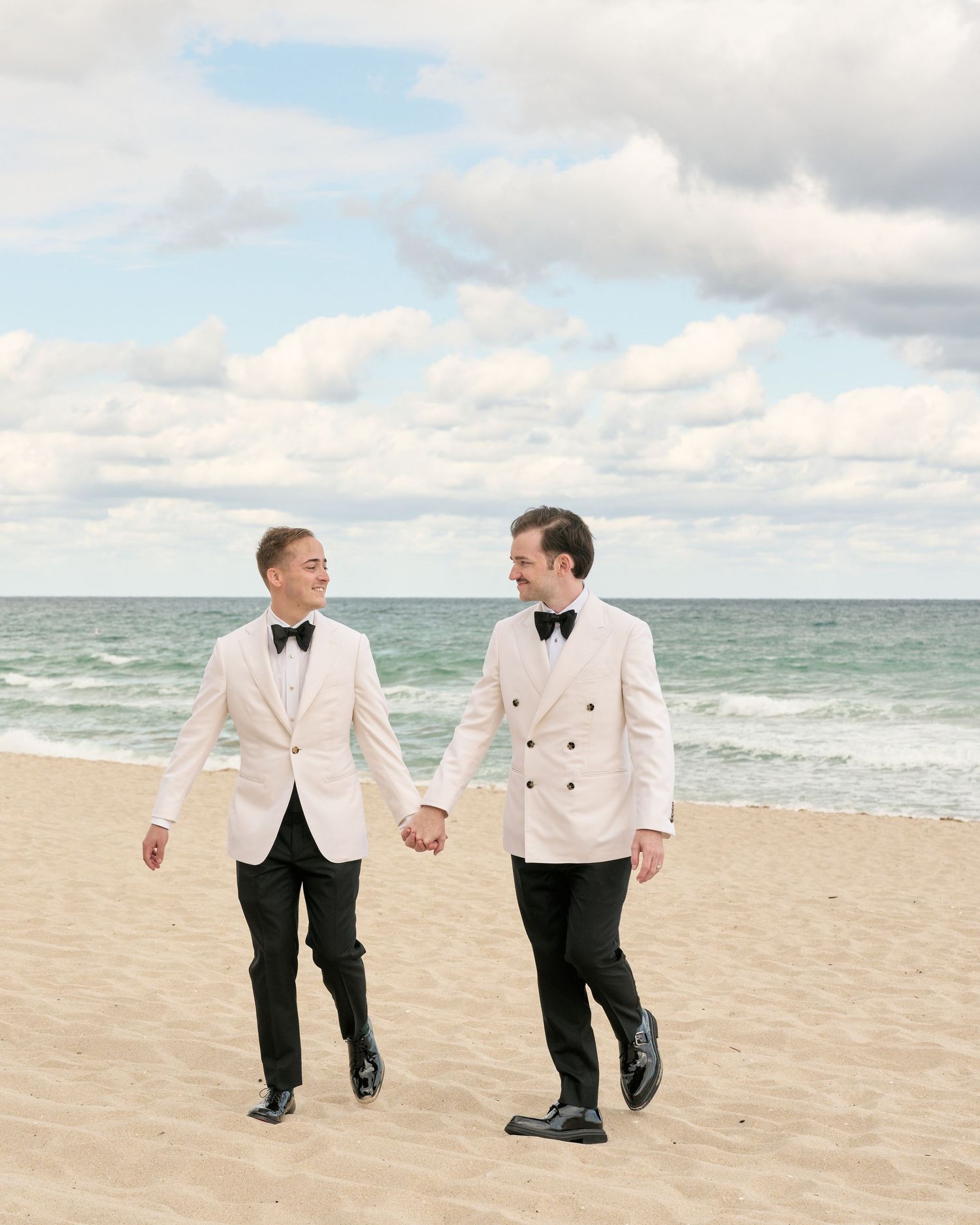 A couple in matching white formal wear walking hand in hand on a sandy beach with ocean waves and cloudy sky backdrop.