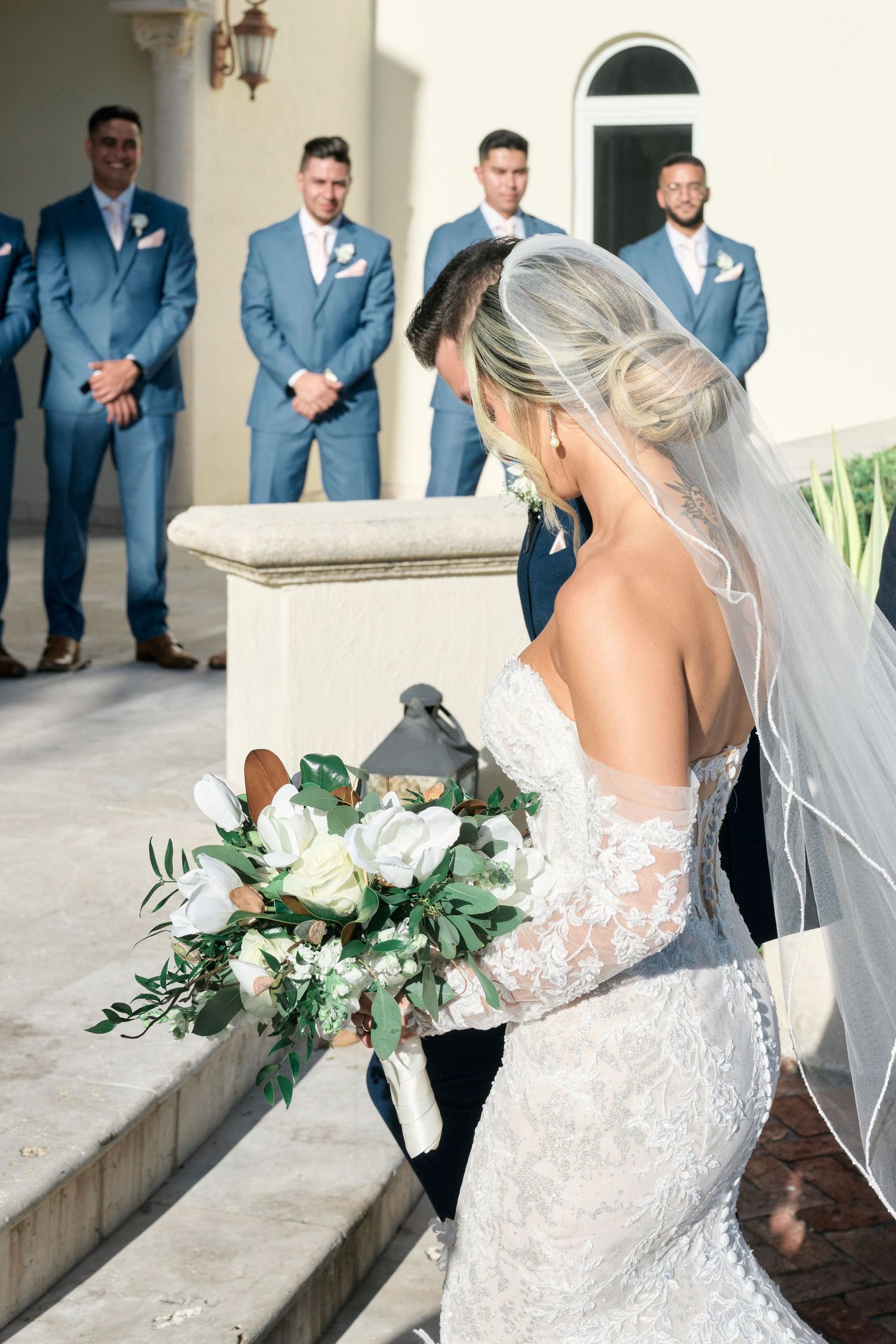 A bride in a lace wedding gown holds a white and green bouquet on stone steps.