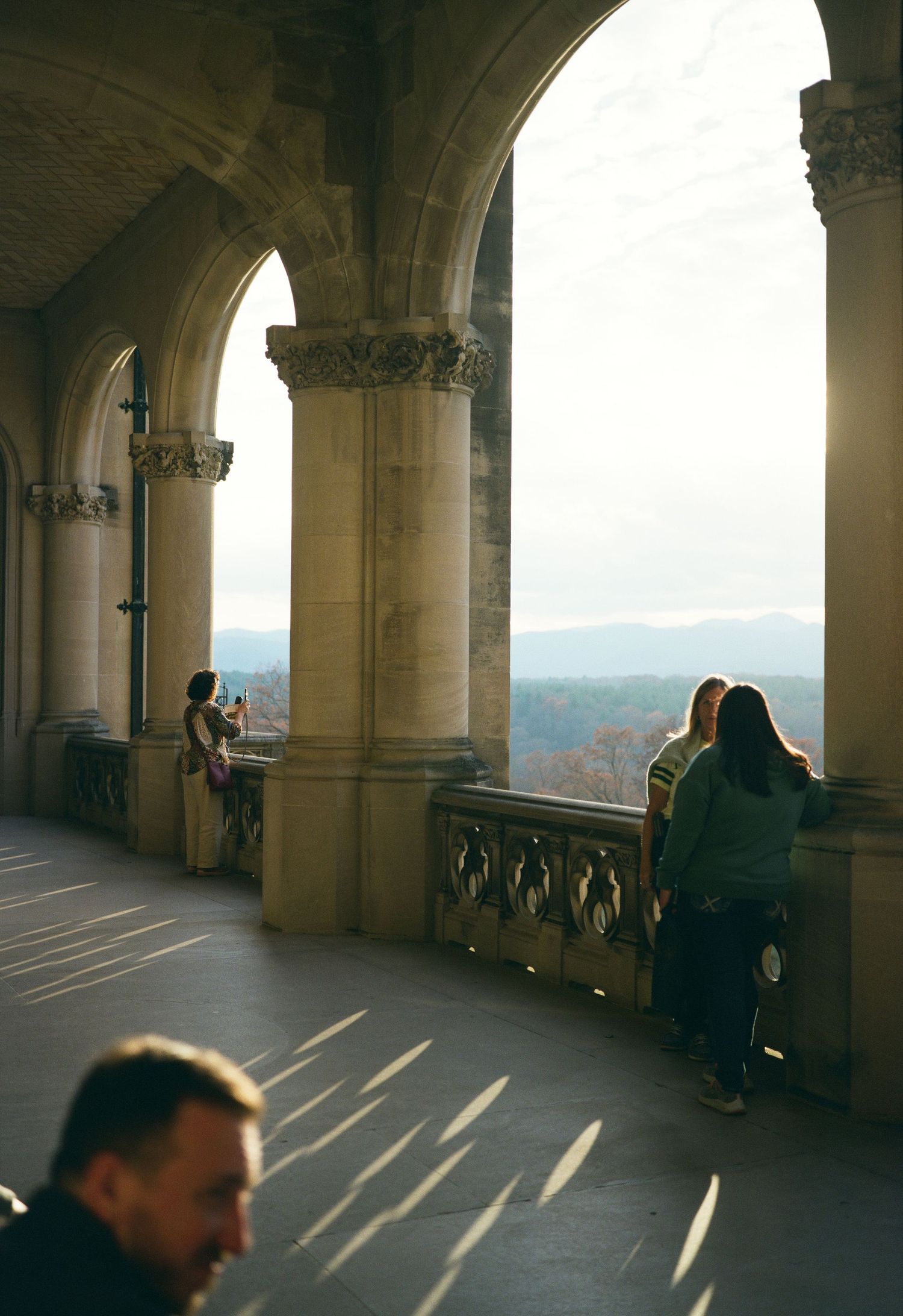 Sun streams through gothic stone arches as visitors enjoy mountain views from castle balcony.