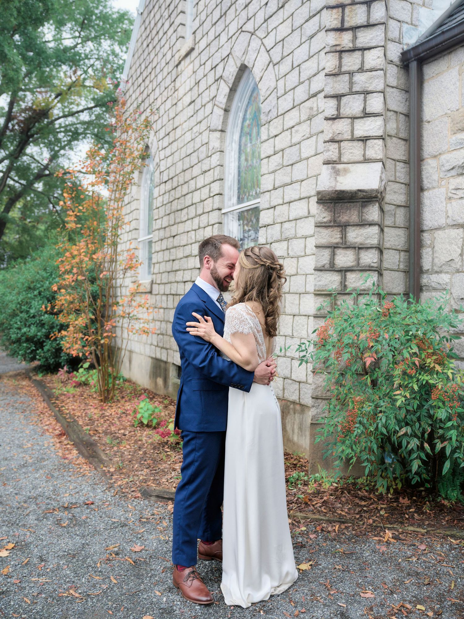 A wedding couple shares an intimate moment against a stone church wall surrounded by autumn foliage.