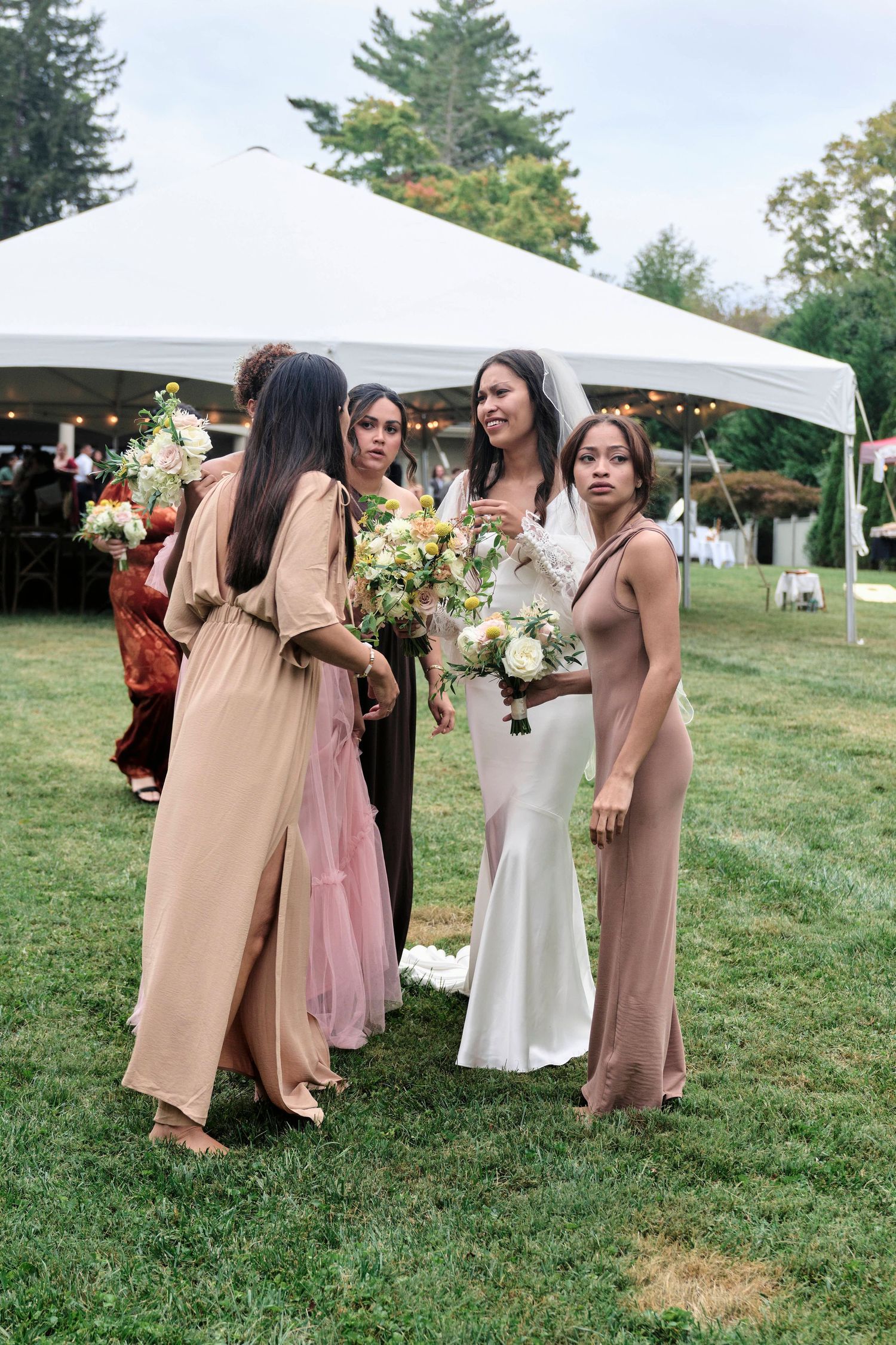Bridal party in neutral dresses gather with bouquets under outdoor tent.