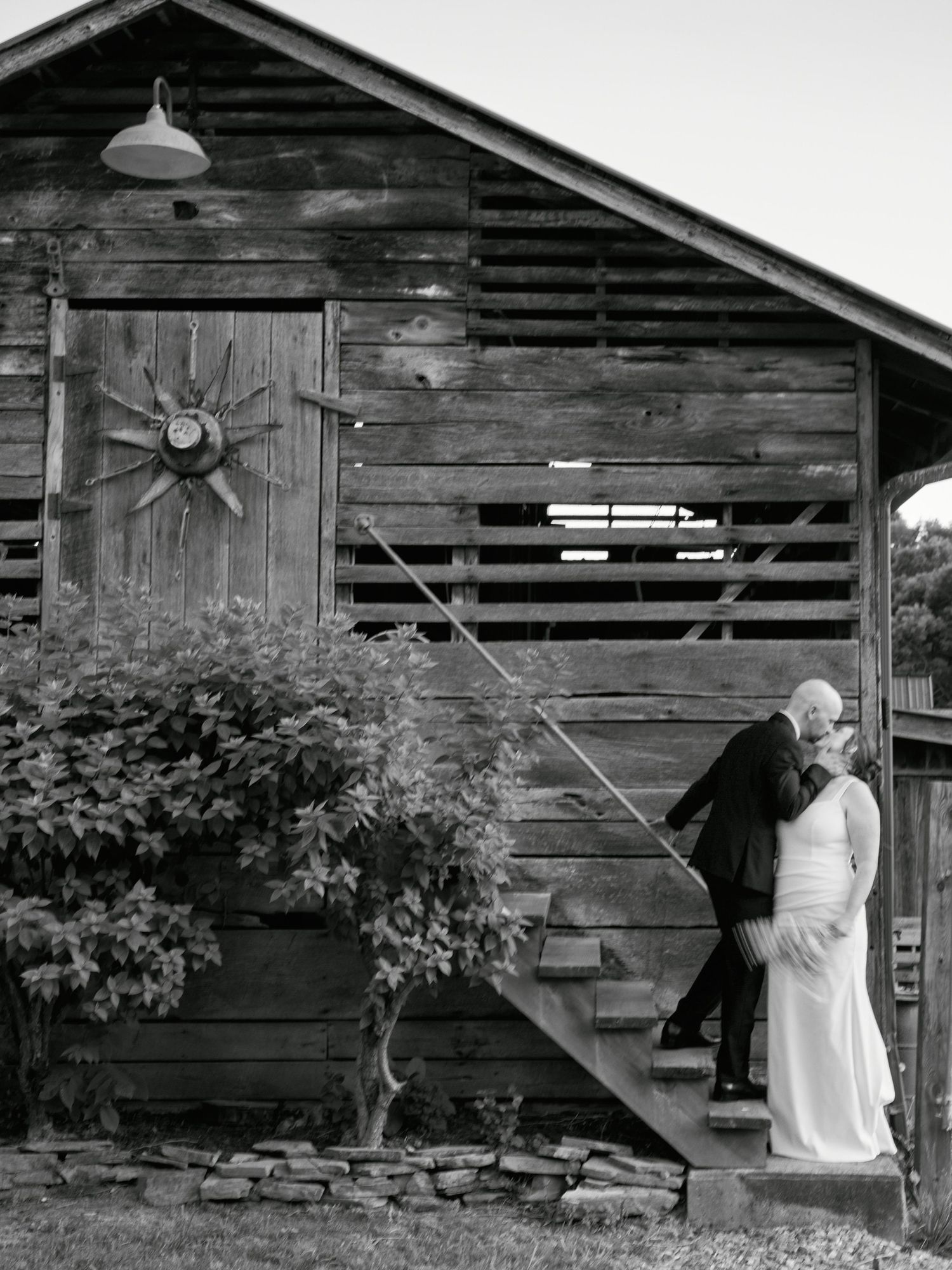 A black and white wedding photo featuring a rustic barn setting with wooden stairs and greenery.