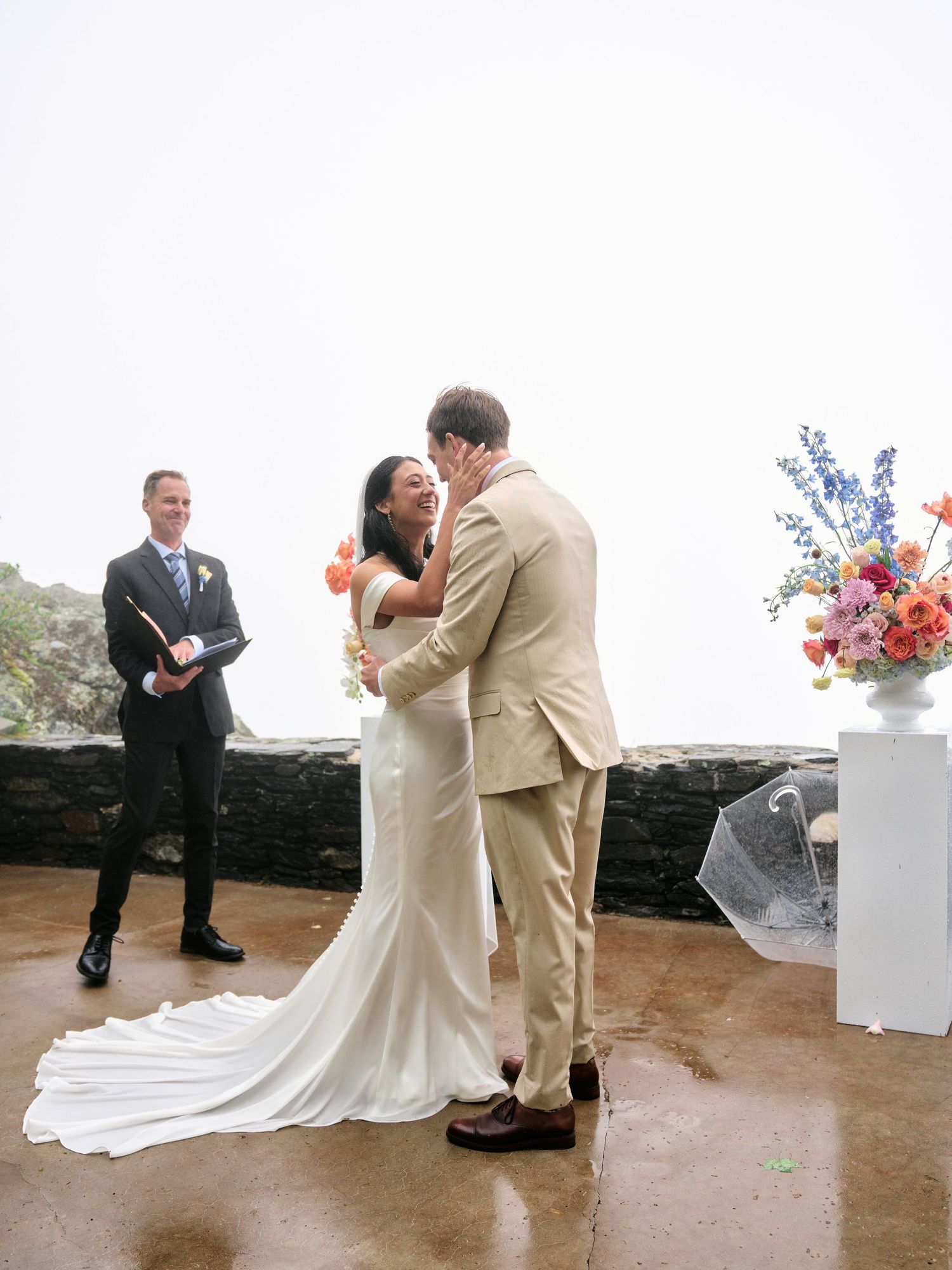 Bride and groom share first kiss at outdoor wedding ceremony with colorful floral arrangement and officiant nearby.