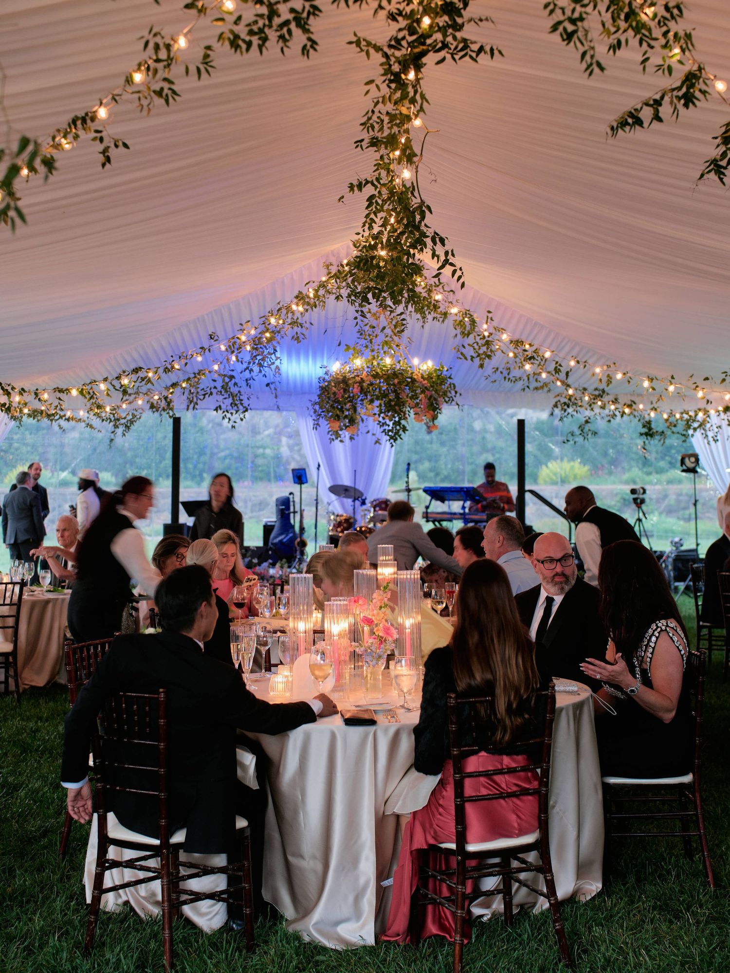 Elegant wedding reception tent with twinkling lights and floral decor as guests dine at round tables under draped fabric ceiling.