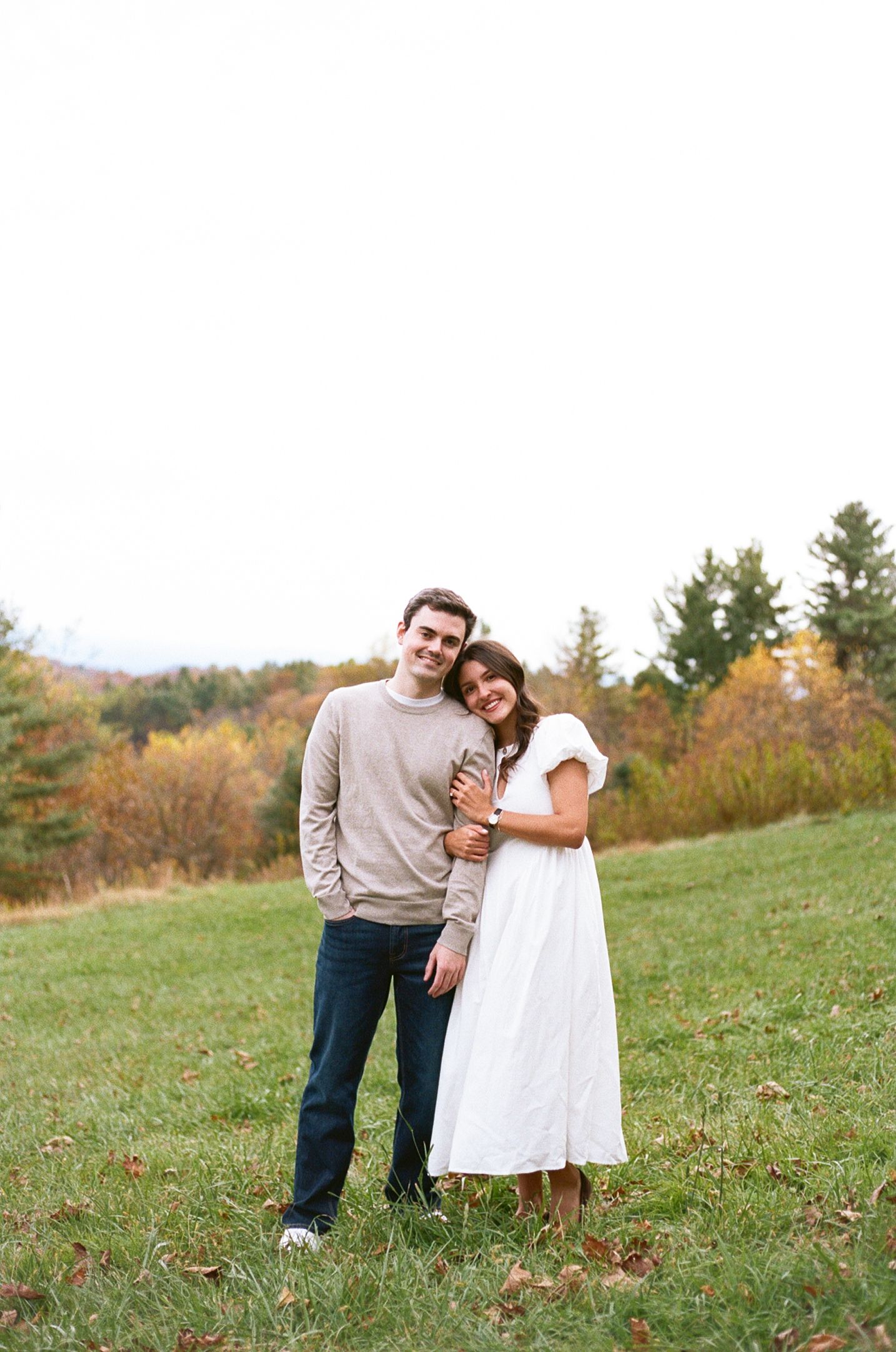 Couple embraces while standing in a grassy autumn field with fall foliage in the background.