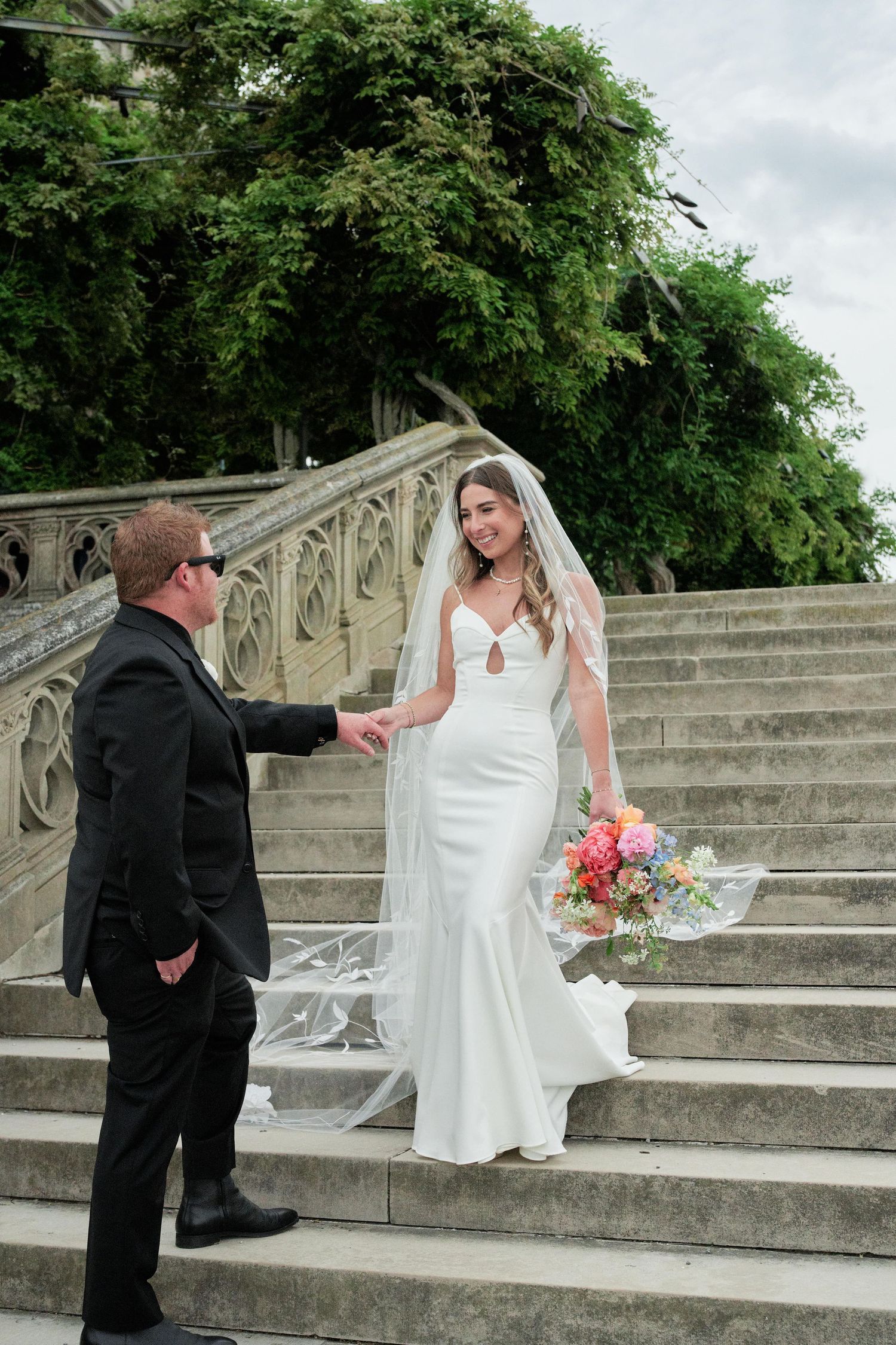 A wedding couple ascends elegant stone steps with ornate railings surrounded by lush green trees in a romantic outdoor setting.