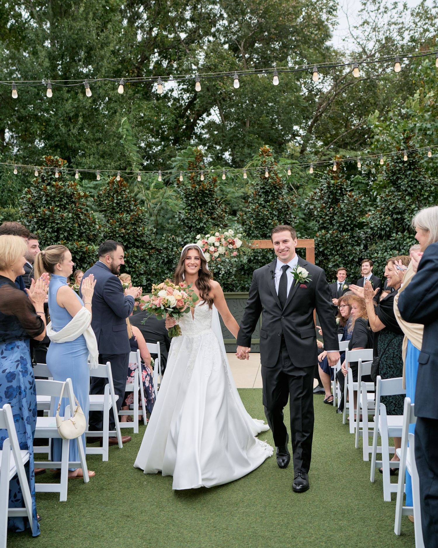 Bride and groom walk down outdoor wedding aisle lined with white chairs and string lights, guests applauding, lush greenery backdrop.