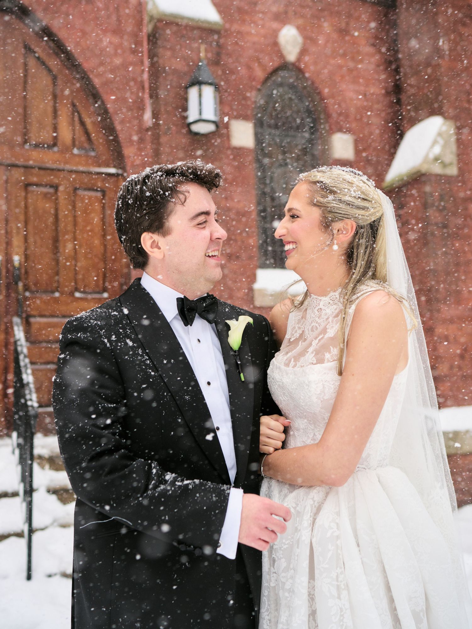Bride and groom smiling at each other in snow outside red brick church, she in lace gown with veil, he in black tuxedo.