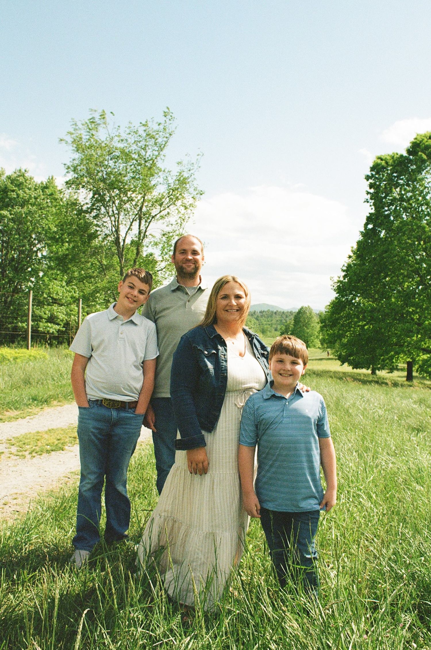 Family of four smiling outdoors in a lush green field with trees and a path on a sunny day.