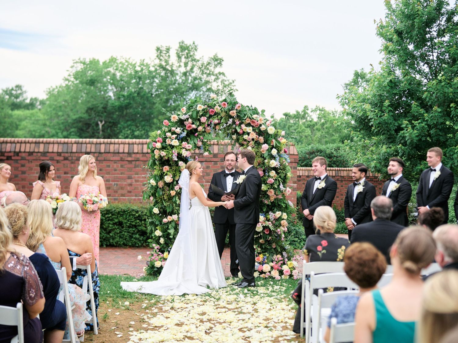 Garden wedding ceremony with rose-covered arch and guests seated on white chairs with flower petals.