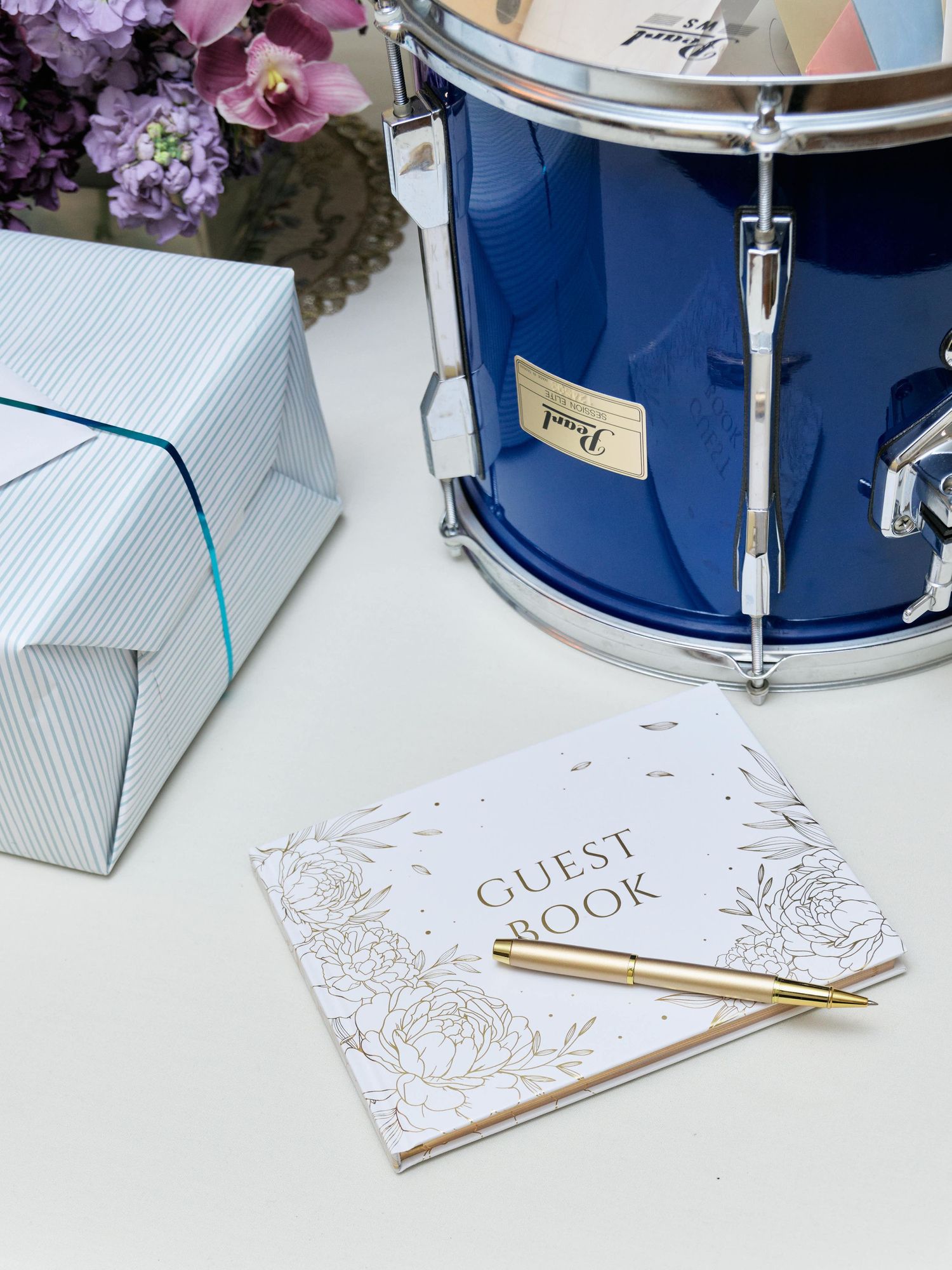Wedding guest book with gold pen displayed next to blue drum and floral decorations.